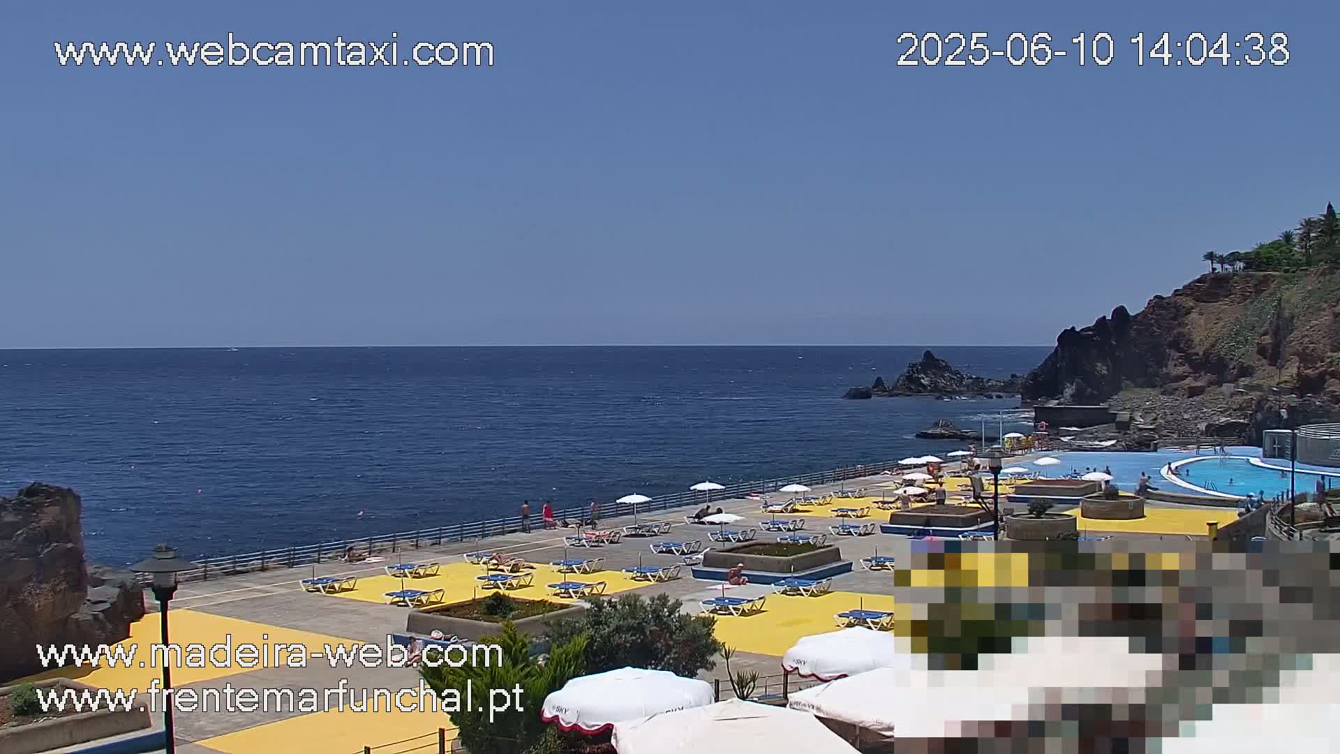 An outdoor swimming pool complex with yellow deck chairs and umbrellas sits on a rocky coast overlooking a calm ocean under a clear blue sky.