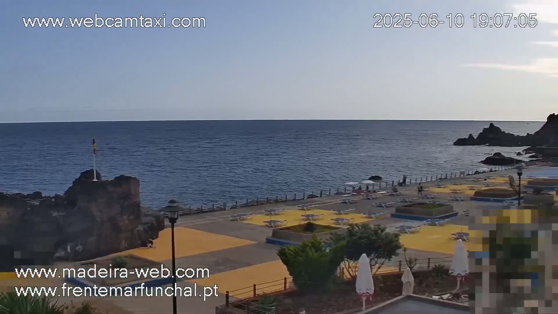 A mostly clear, twilight sky overlooks a rocky coast with a sunbathing area featuring yellow-striped lounge chairs and umbrellas.