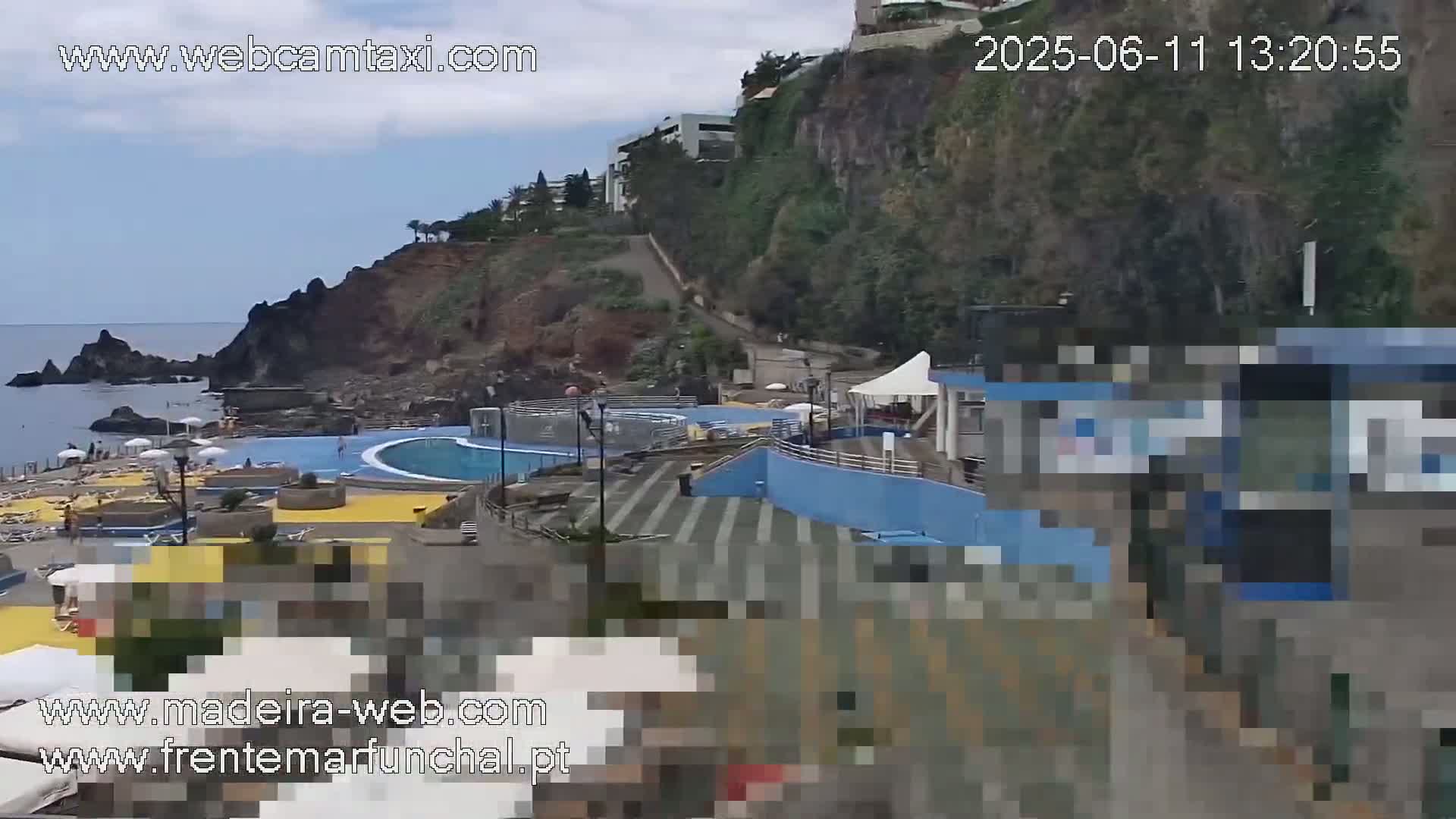 An outdoor scene shows a partially-cloudy day overlooking a seaside pool and beach area nestled against a rocky cliffside with buildings perched atop.