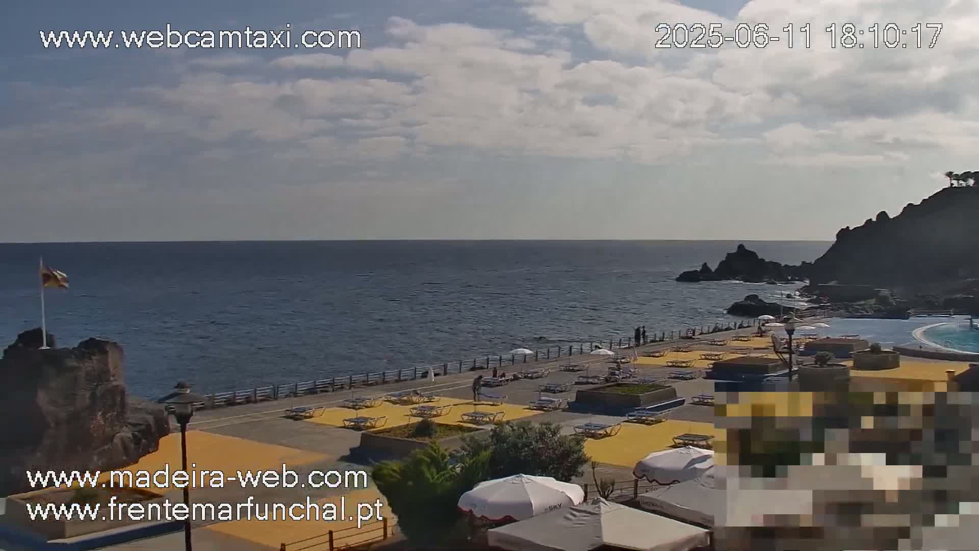 A mostly sunny outdoor scene shows a paved seaside area with yellow lounge chairs and umbrellas, a dark rocky coastline, and calm blue ocean water.