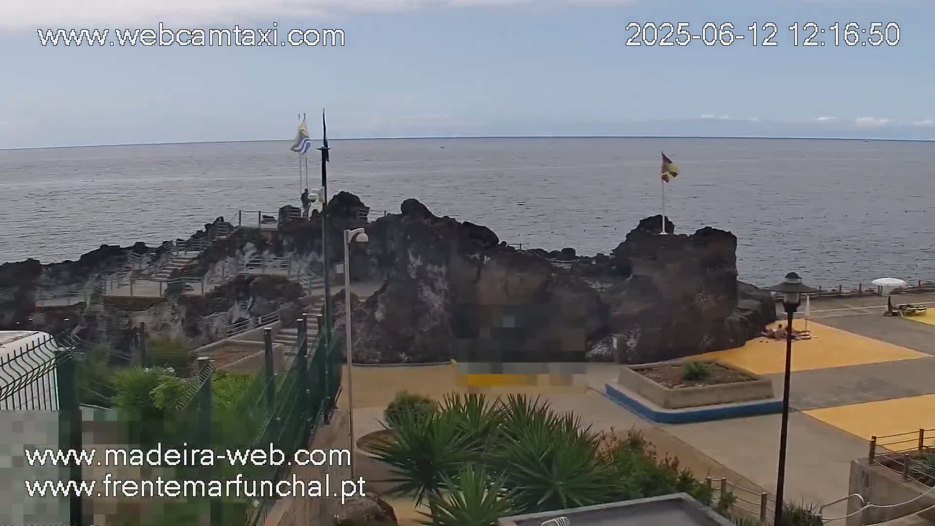 A rocky outcrop juts into a calm ocean under a mostly cloudy sky, with a paved area and sparse vegetation nearby.