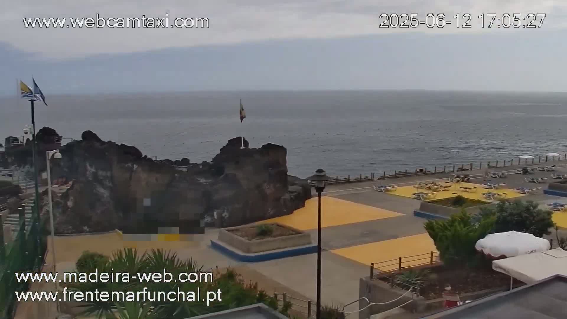An overcast day reveals a coastal scene with a rocky outcrop, a paved area with yellow sections and lounge chairs, and a calm ocean stretching to the horizon.