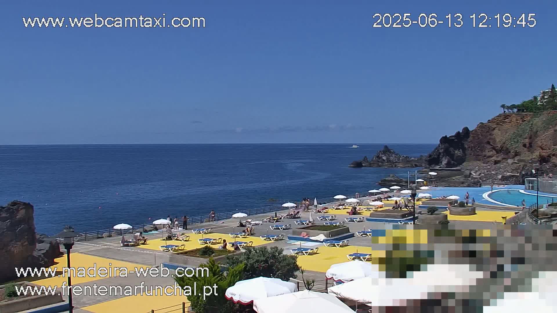 A sunny outdoor scene shows a seaside pool and patio area with numerous people lounging under umbrellas, all situated against a backdrop of dark volcanic rock cliffs and a calm blue ocean.