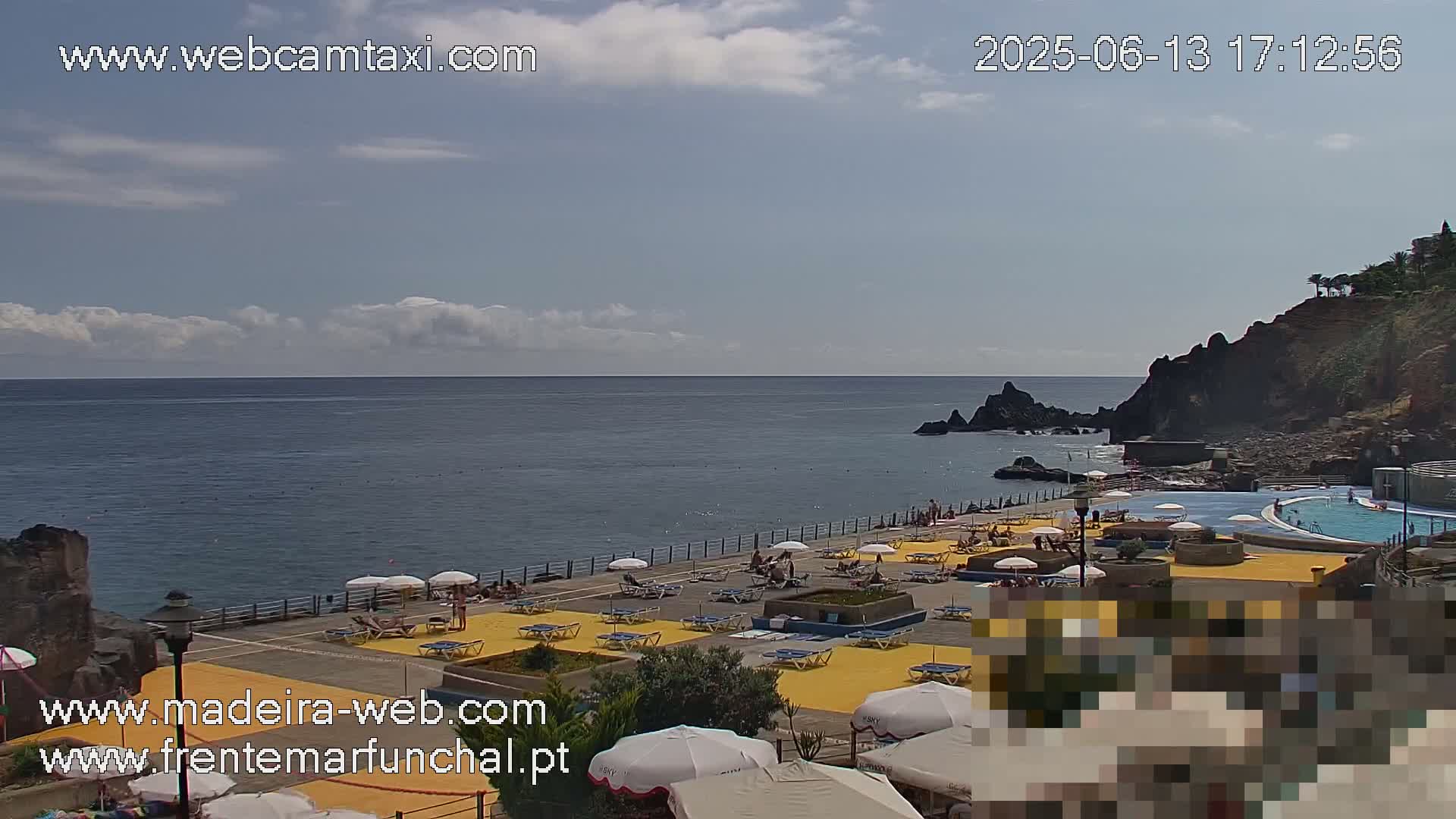 A partly sunny outdoor scene shows a seaside area with a swimming pool and sunbathers on yellow lounge chairs, all situated on a paved area next to rocky cliffs and a calm ocean.