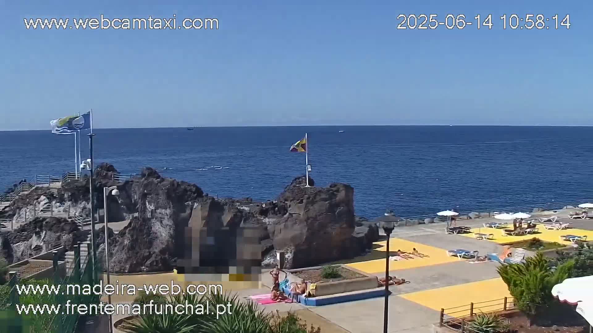 A sunny day at an oceanside outdoor area with dark volcanic rock formations, a paved sunbathing area with people sunbathing and lounging, and flags on poles.