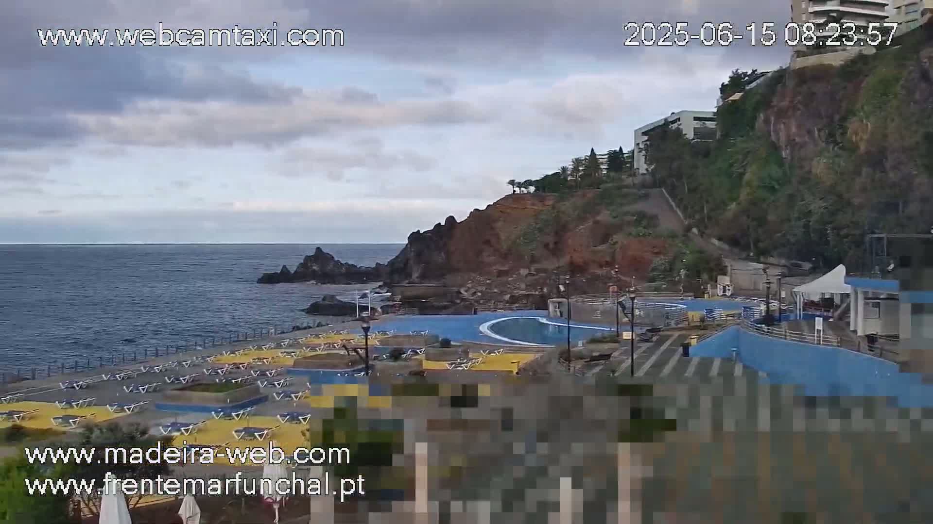 An outdoor scene shows a coastline with a swimming pool complex, a rocky cliff, and a calm ocean under a mostly cloudy sky.