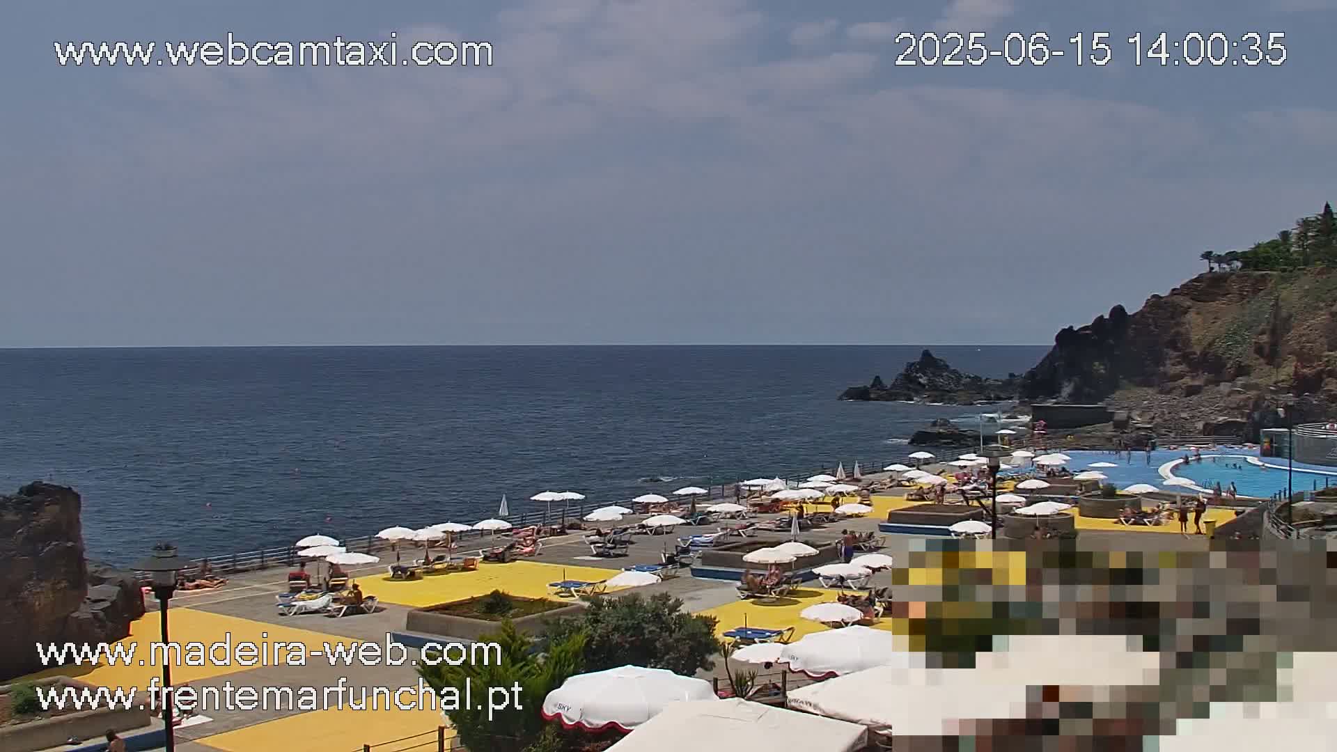 A seaside outdoor pool and sunbathing area with many umbrellas and people are visible on a partly cloudy day.