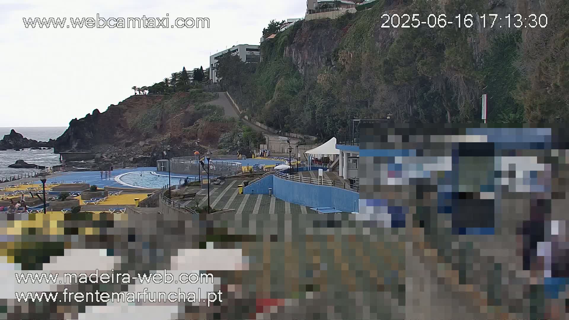 An outdoor scene on an overcast day shows a coastal swimming pool complex situated below a cliffside with buildings and vegetation.