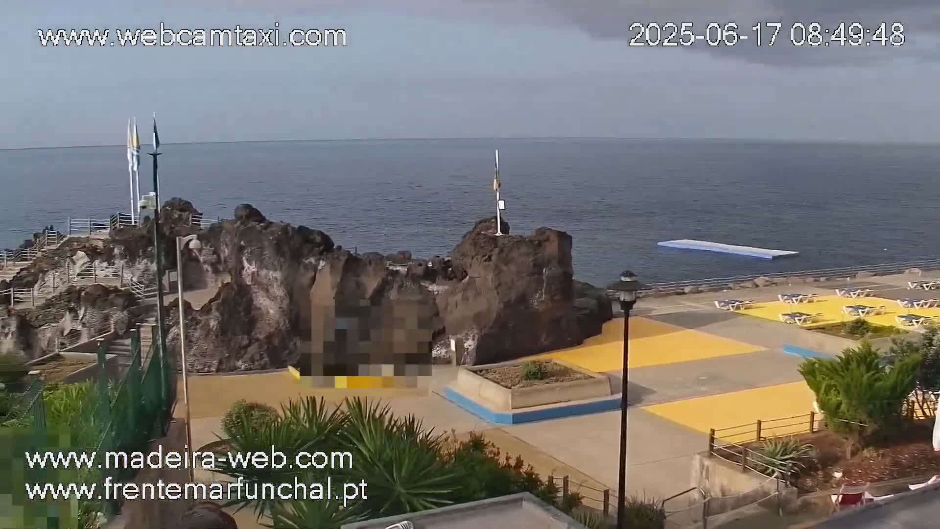 A rocky coastal area with a paved plaza containing yellow sections, a floating platform in the ocean, and overcast skies.