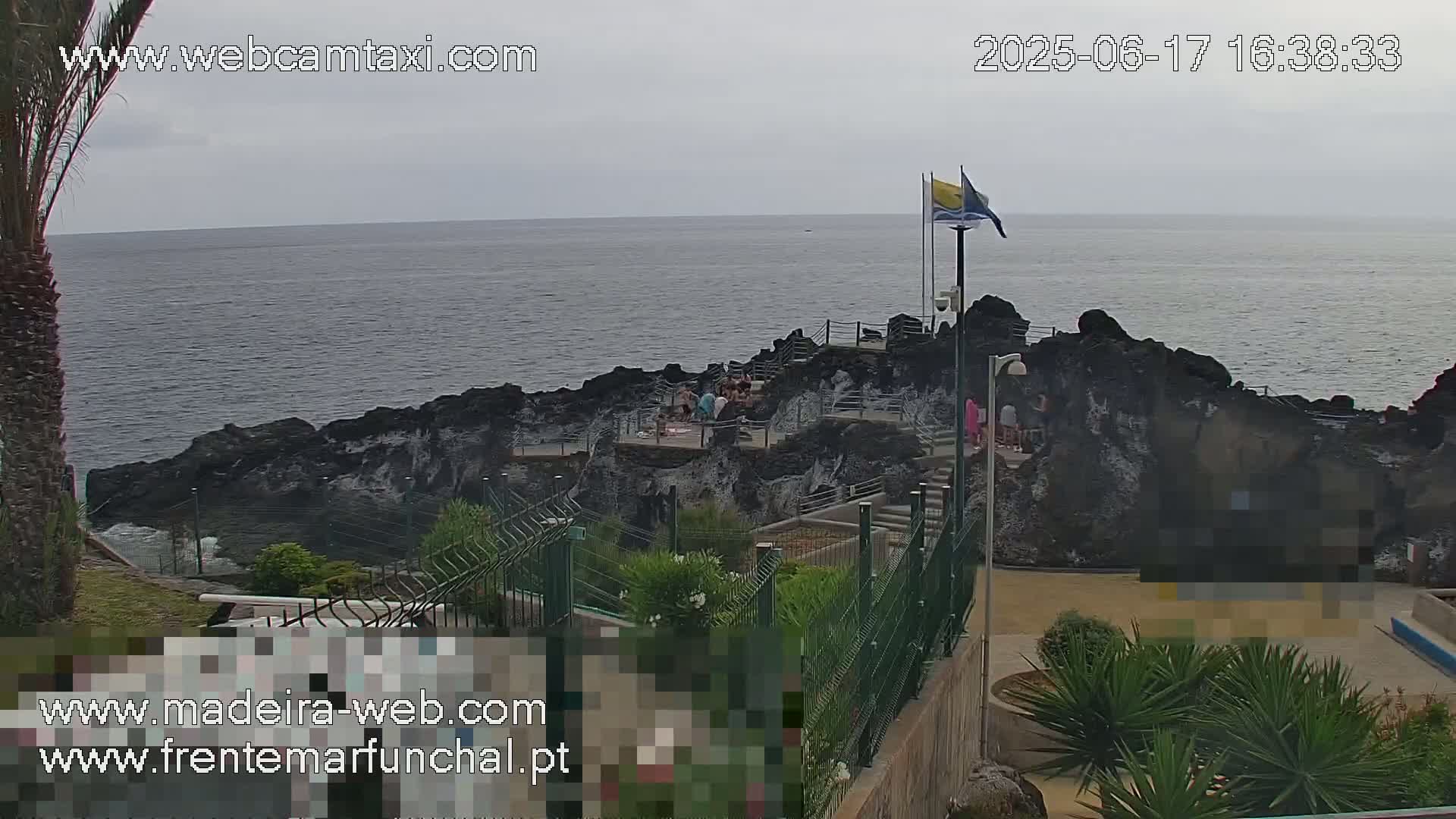 A rocky outcrop juts into a calm, gray ocean under an overcast sky, with a few people gathered on the rocks and surrounding landscaped areas.