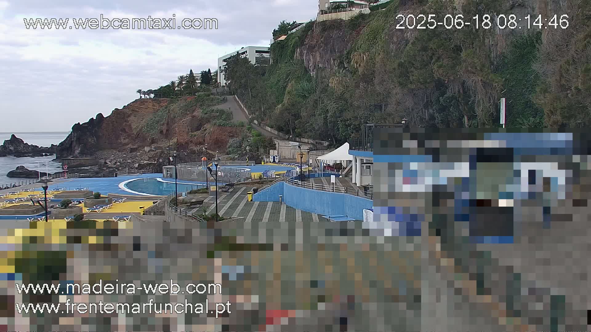 An outdoor pool complex with surrounding buildings and a rocky coastline is visible under an overcast sky.
