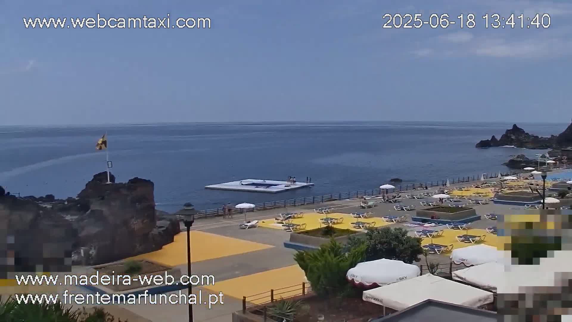 A sunny day overlooking an oceanside resort with a floating platform, numerous lounge chairs under umbrellas, and a rocky coastline.