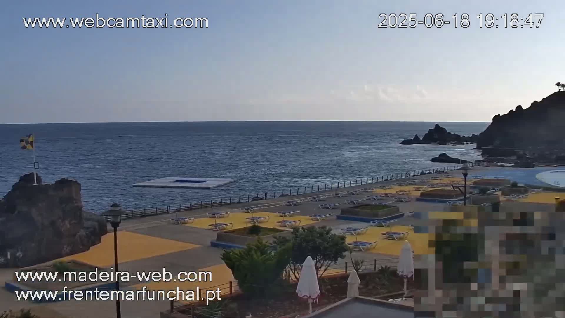 A mostly clear, daytime view of a coastal area with a calm ocean, a paved walkway lined with lounge chairs, and dark volcanic rock formations.