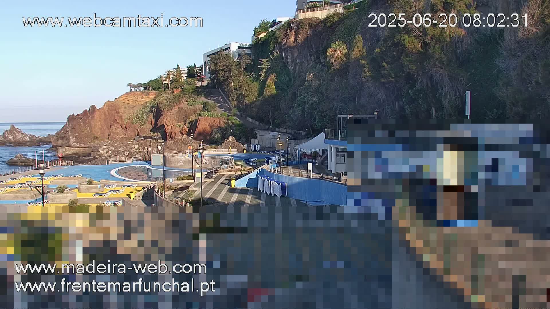 An outdoor oceanside swimming pool area with a rocky cliff behind it, on a clear, sunny morning.