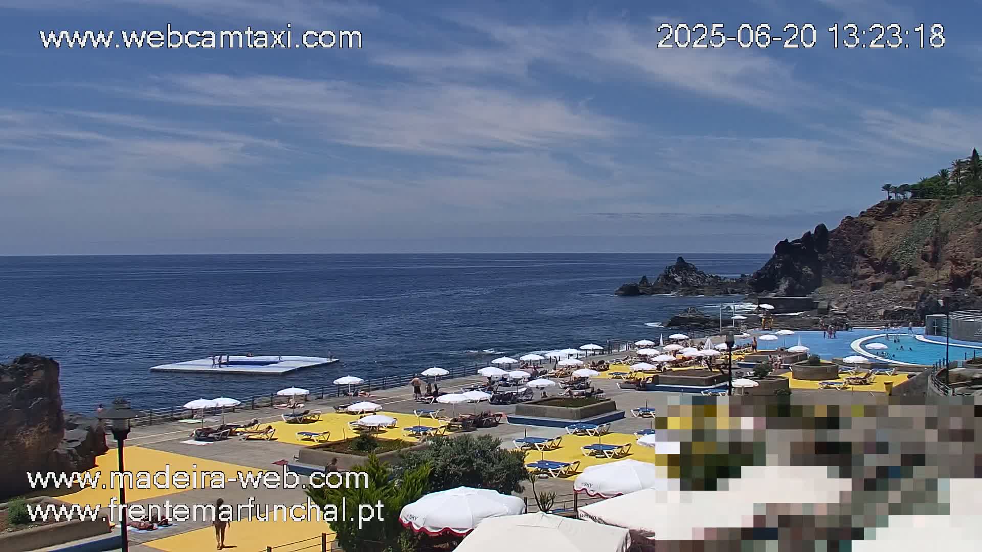 An outdoor scene shows a partly sunny day with a mostly clear sky over a rocky coastline, featuring a swimming pool complex with numerous yellow umbrellas and people enjoying the water and sun.