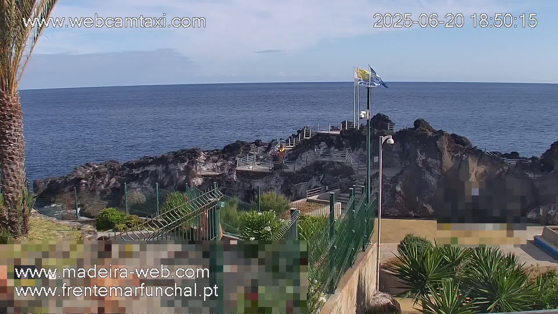 A rocky coastal area with a walkway and flags overlooks a calm ocean under a clear sky.