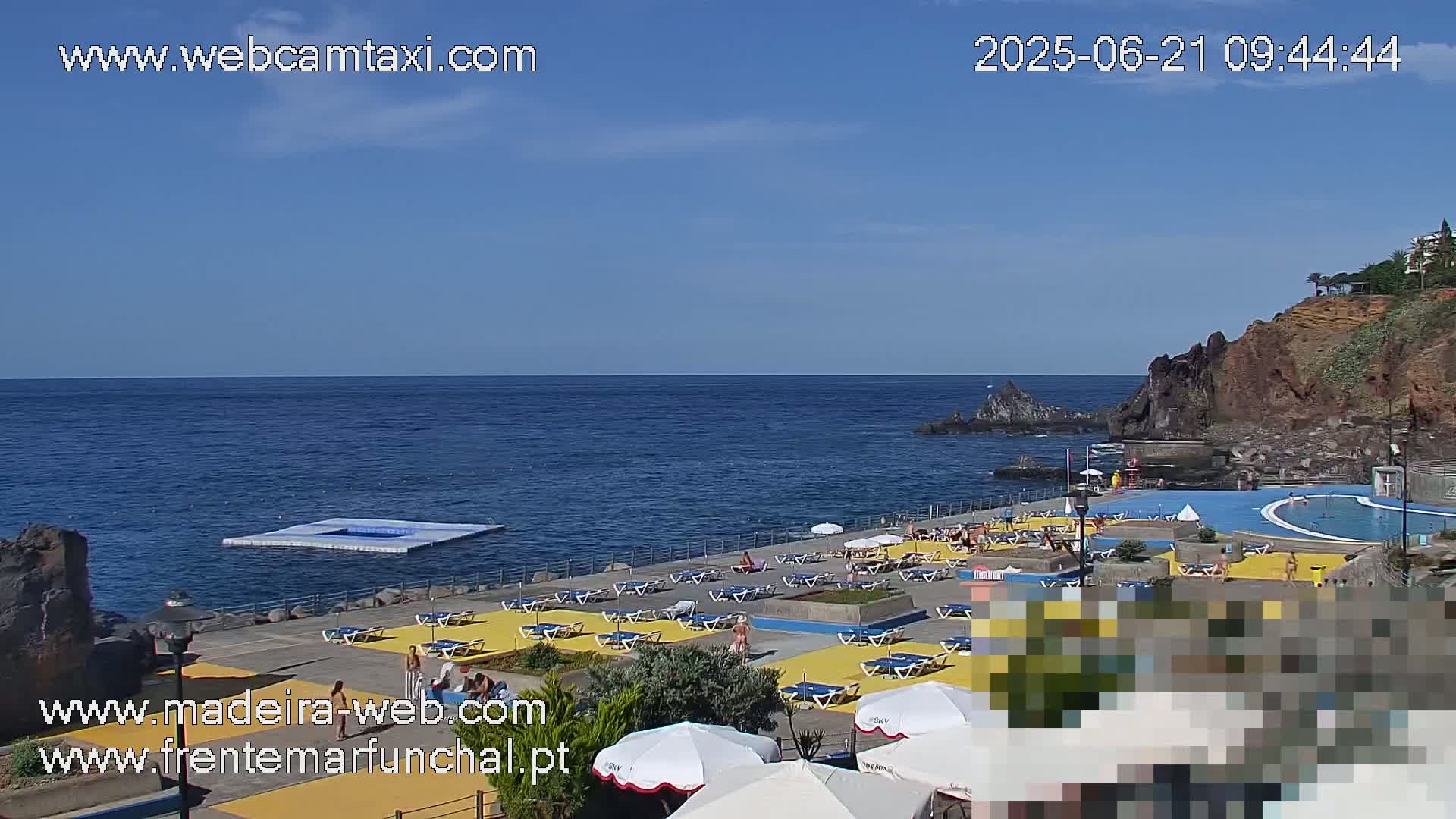 An outdoor swimming pool and sunbathing area are situated on a rocky coast overlooking a calm ocean under a clear blue sky.