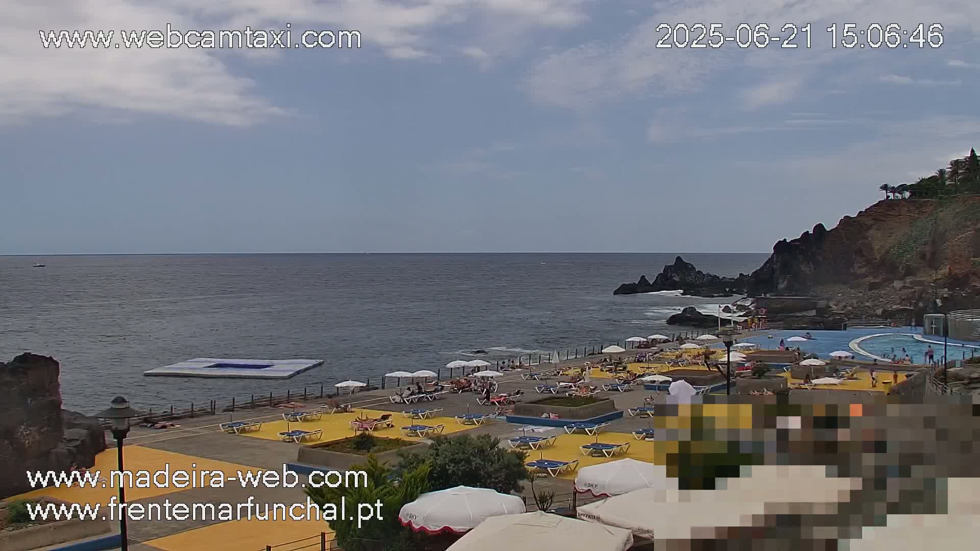A partly cloudy day reveals an oceanside scene with a swimming pool and sunbathers relaxing under umbrellas on a yellow-tiled deck near rocky cliffs.