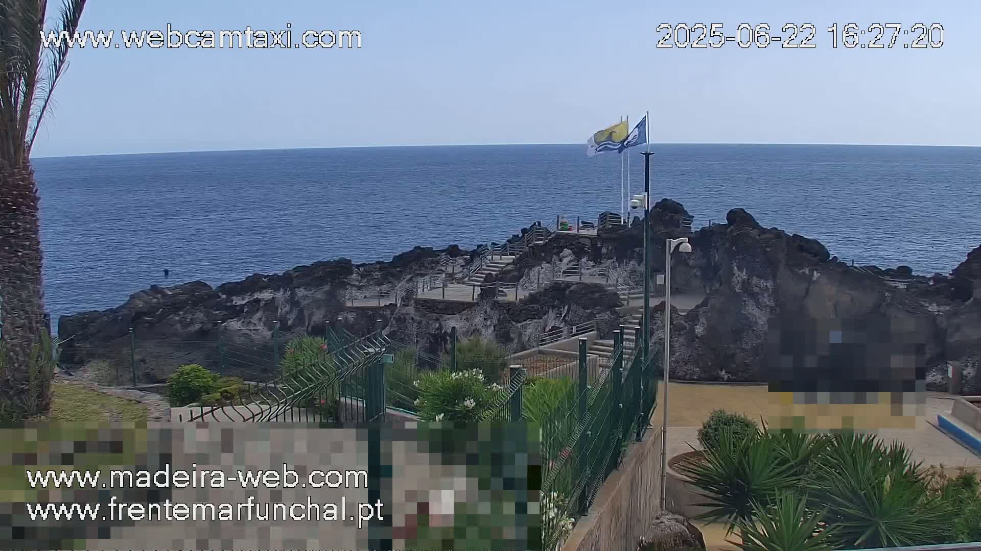 A rocky coast with stairs leading down to the water, under a clear blue sky, is partially enclosed by a green metal fence and surrounded by low-lying vegetation.