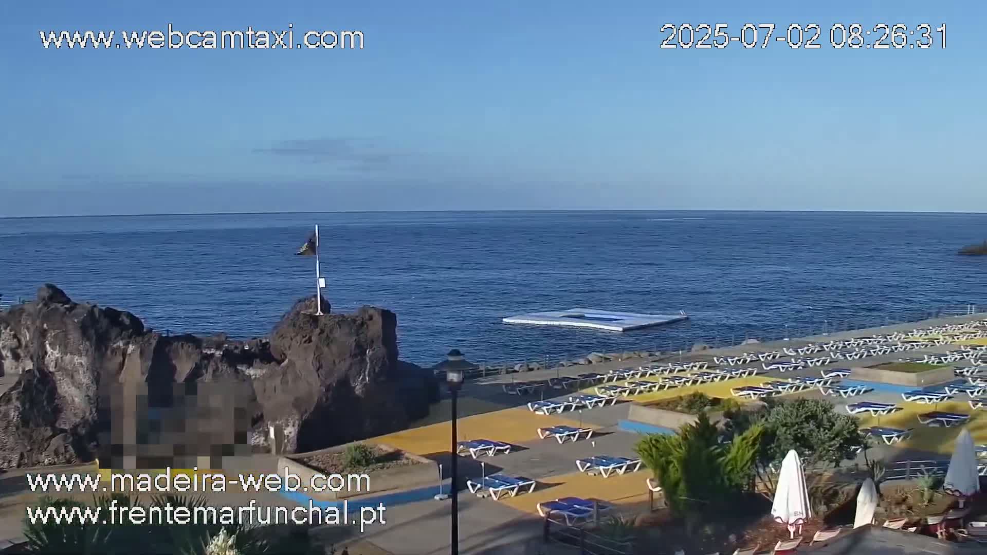 A sunny coastal scene shows a rocky outcrop, a calm ocean, and a sun deck with numerous lounge chairs.