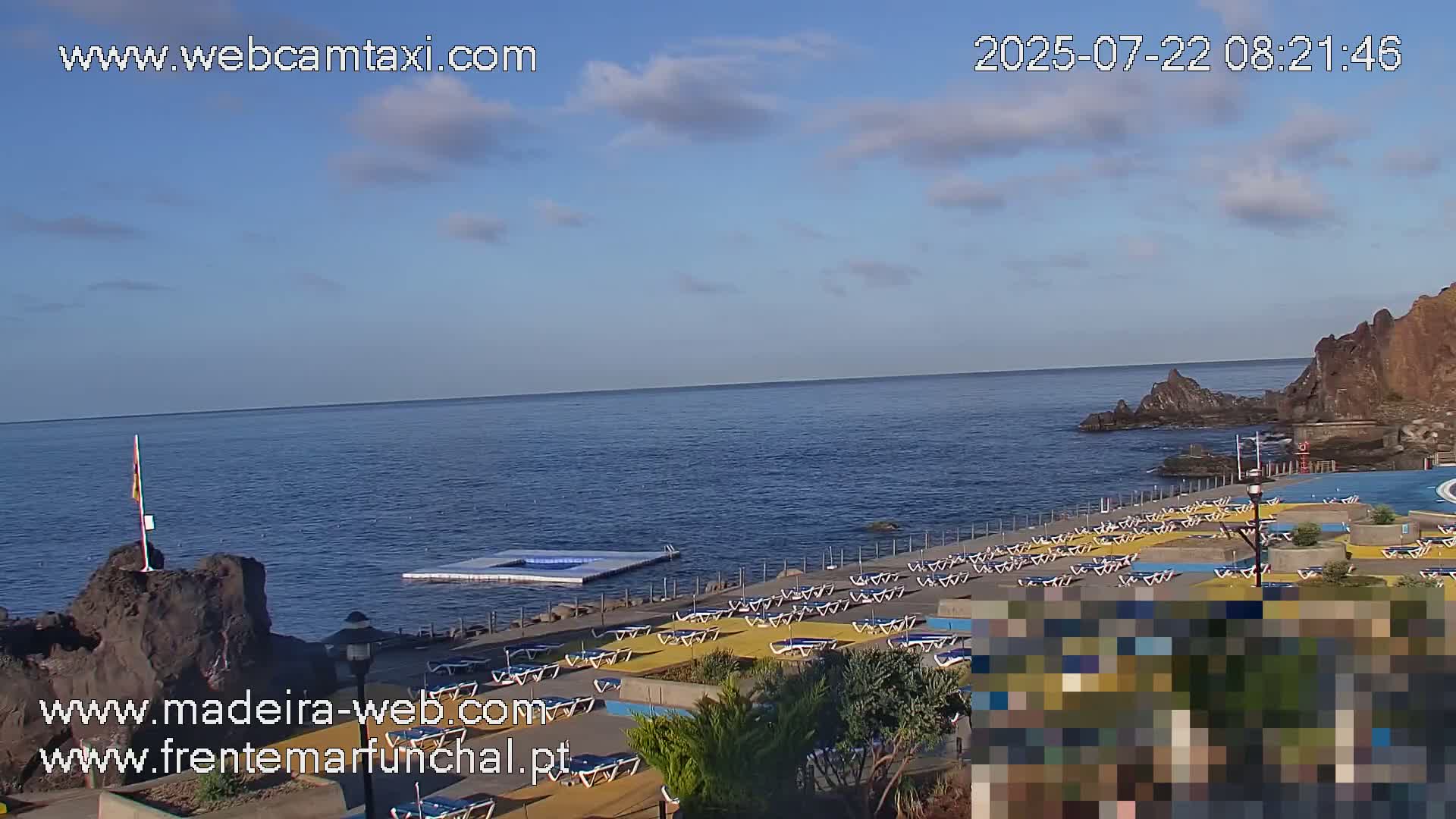 An oceanfront pool area with numerous empty lounge chairs sits under a mostly clear sky with a few scattered clouds.