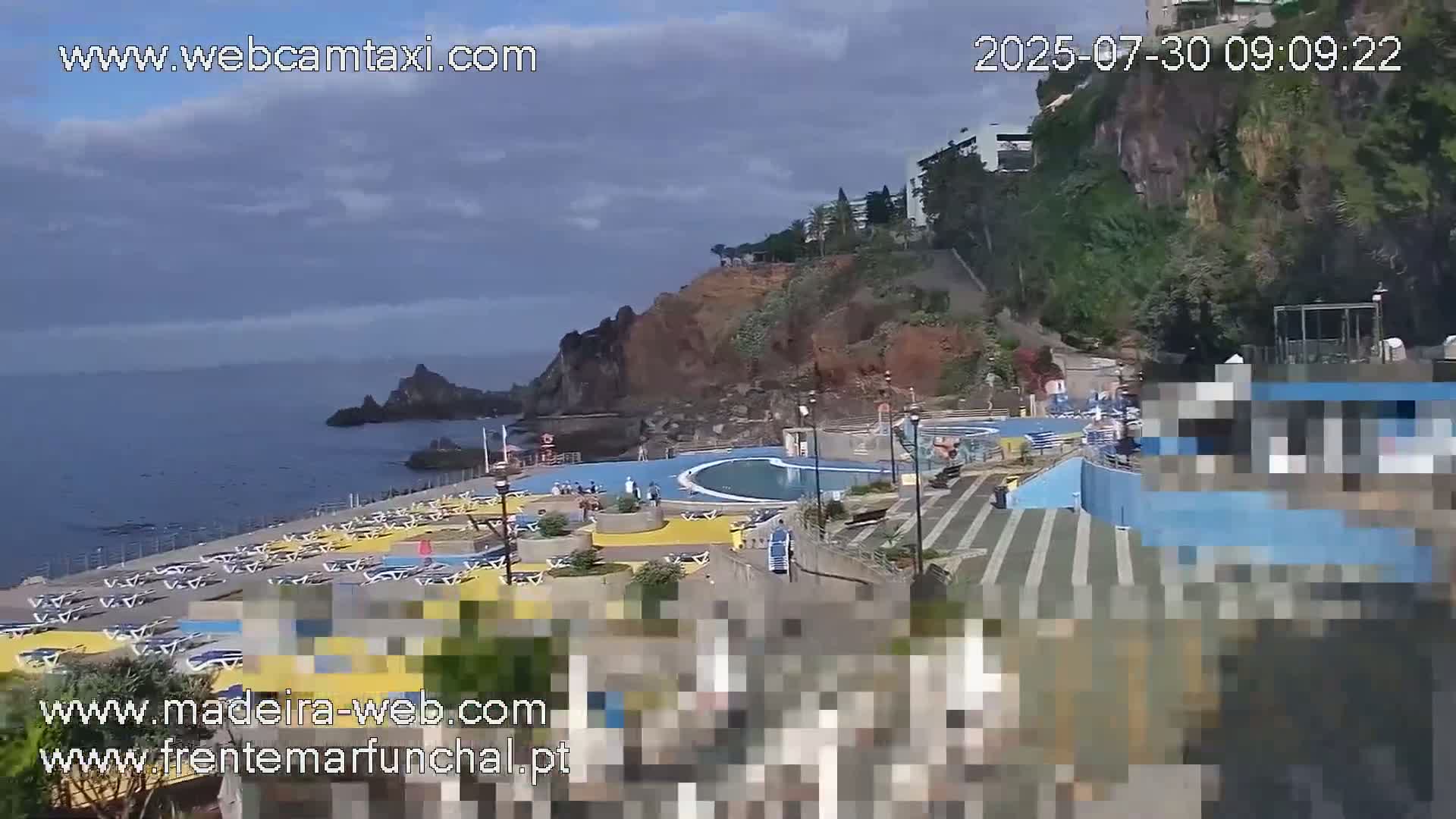 A seaside pool area with many lounge chairs and a partially cloudy sky overlooks a rocky coast and ocean.