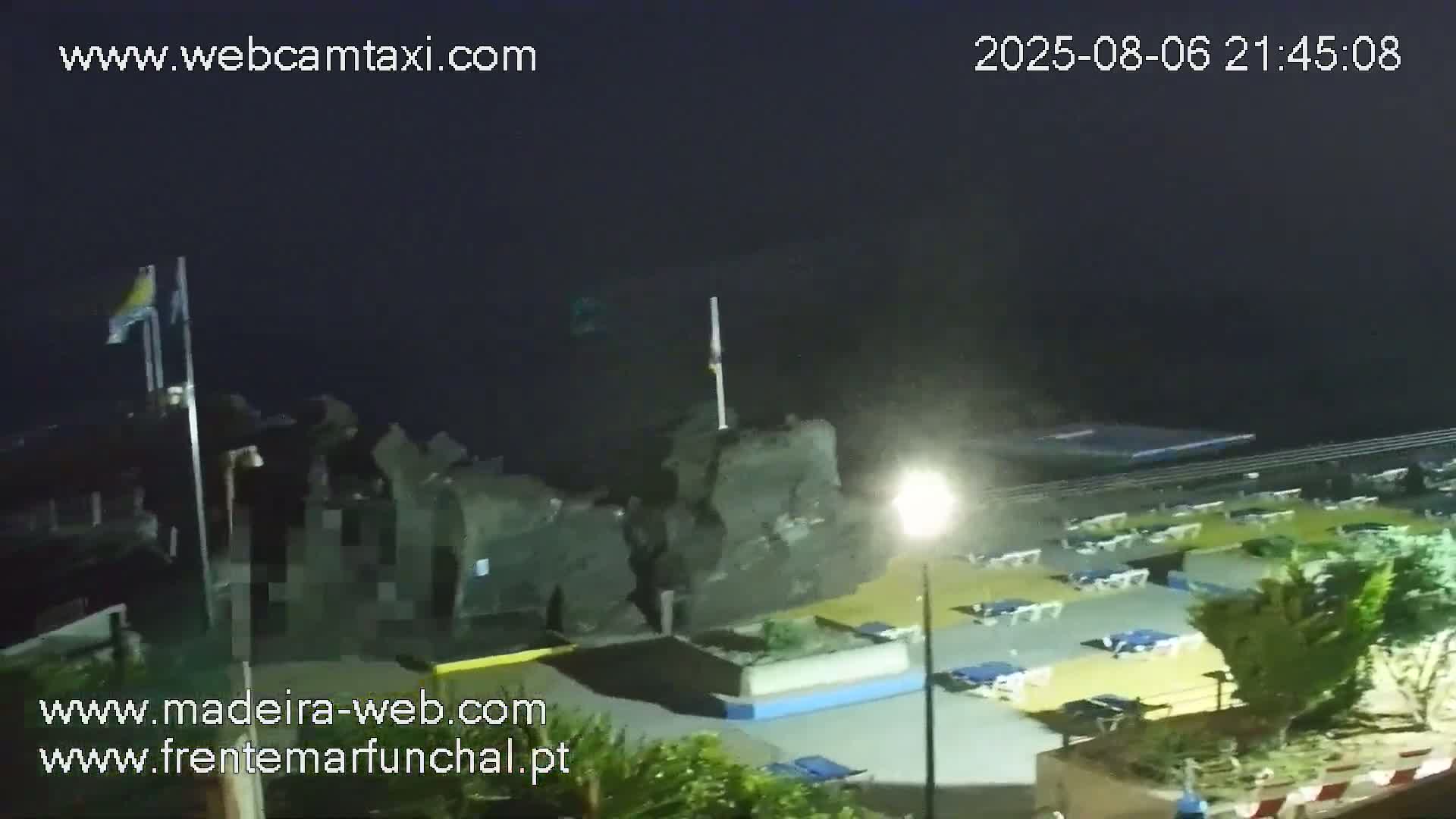 A nighttime view shows a dark, rocky coastline with a beach area containing lounge chairs, illuminated by a lamppost, under a mostly clear, dark sky.