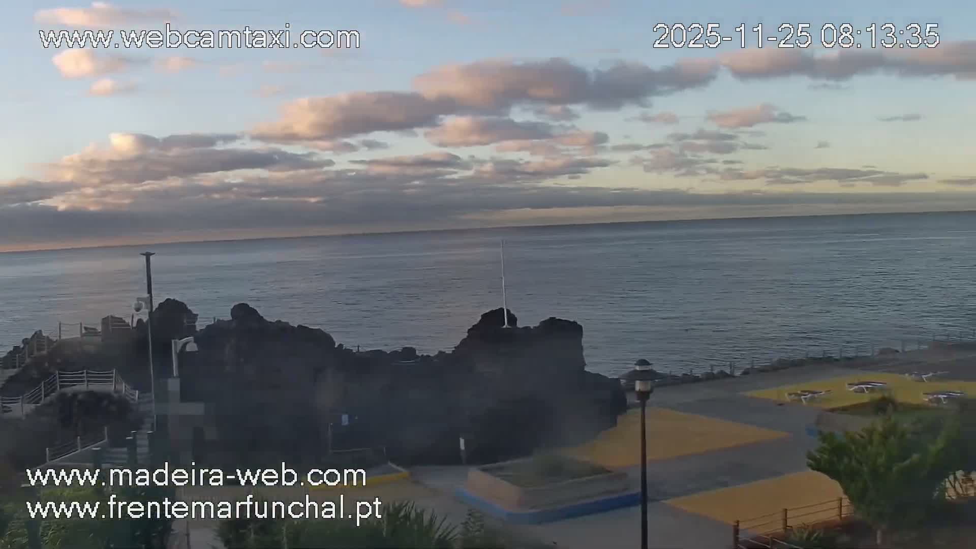 An early morning coastal scene features a calm ocean extending to the horizon under a partly cloudy sky, with rocky outcrops and a paved promenade lined with sun loungers along the shore.