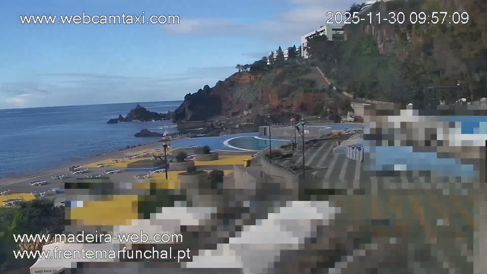 A sunny, partly cloudy day reveals a coastal scene featuring a dark blue ocean meeting a rocky beach lined with sun loungers, adjacent to a modern lido with a blue swimming pool, all overlooked by a steep, green cliff topped with buildings.