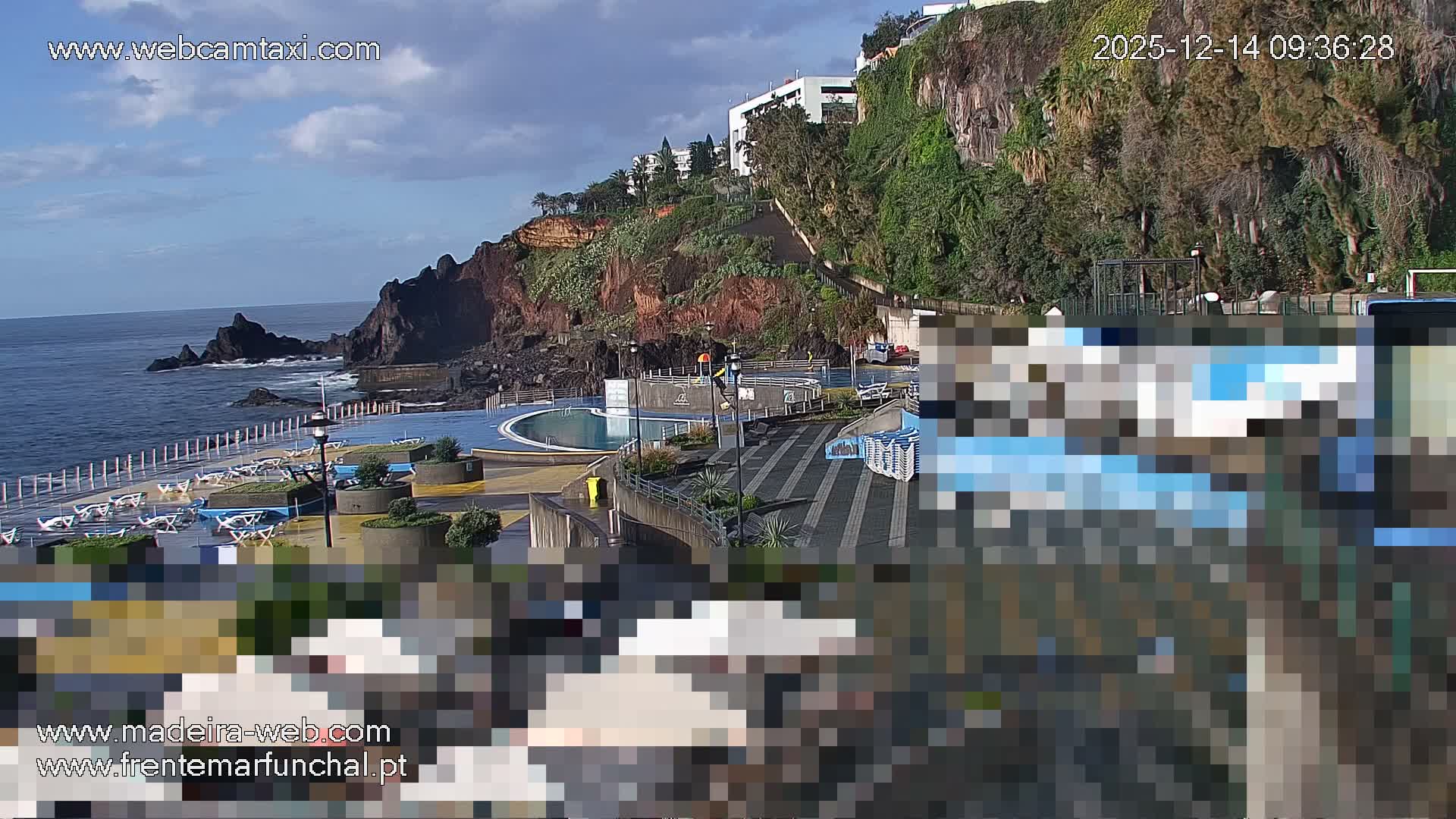 A sunny, partly cloudy day reveals a coastal scene featuring a dark blue ocean meeting a rocky beach lined with sun loungers, adjacent to a modern lido with a blue swimming pool, all overlooked by a steep, green cliff topped with buildings.