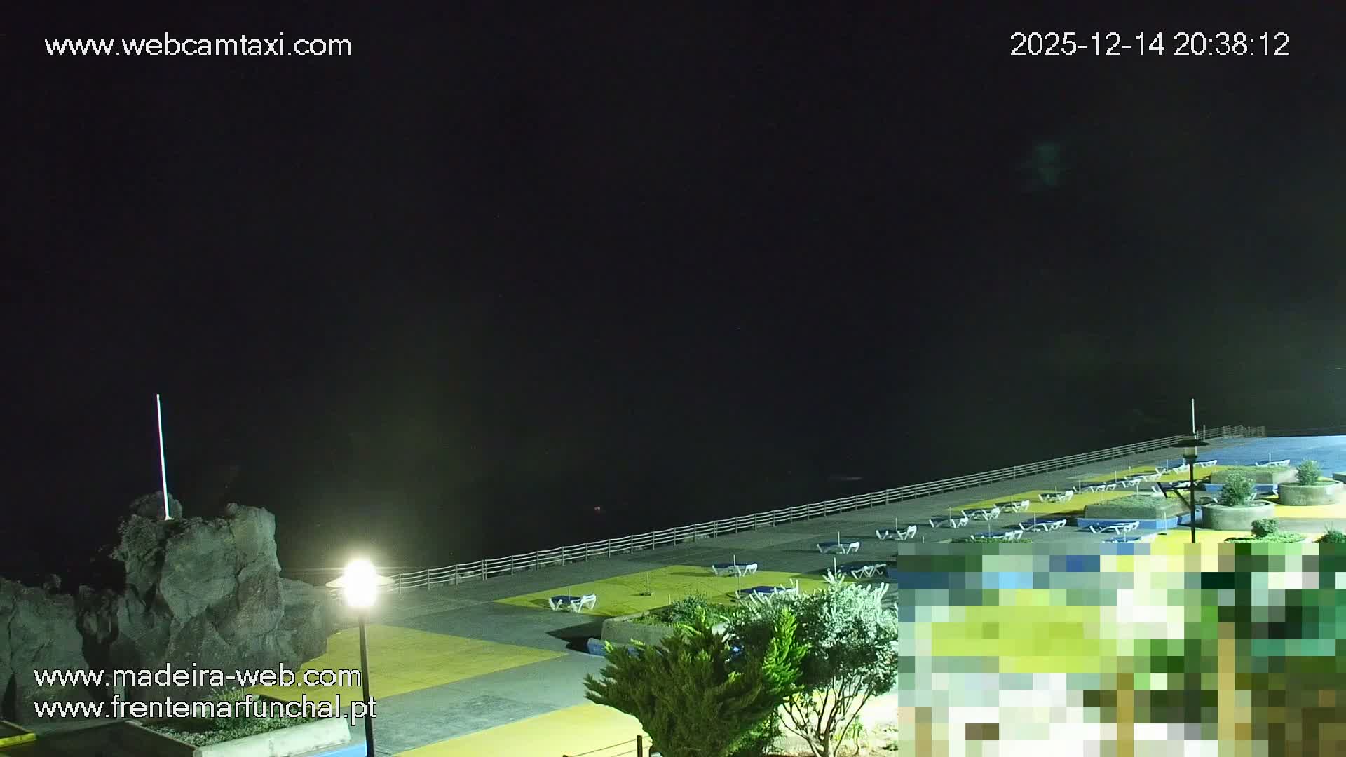 A sunny, partly cloudy day reveals a coastal scene featuring a dark blue ocean meeting a rocky beach lined with sun loungers, adjacent to a modern lido with a blue swimming pool, all overlooked by a steep, green cliff topped with buildings.