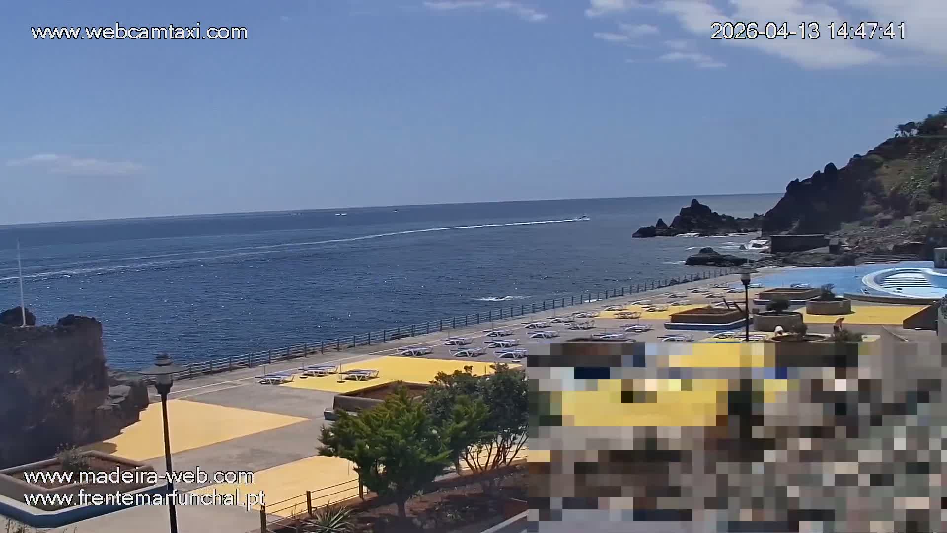 A sunny, partly cloudy day reveals a coastal scene featuring a dark blue ocean meeting a rocky beach lined with sun loungers, adjacent to a modern lido with a blue swimming pool, all overlooked by a steep, green cliff topped with buildings.