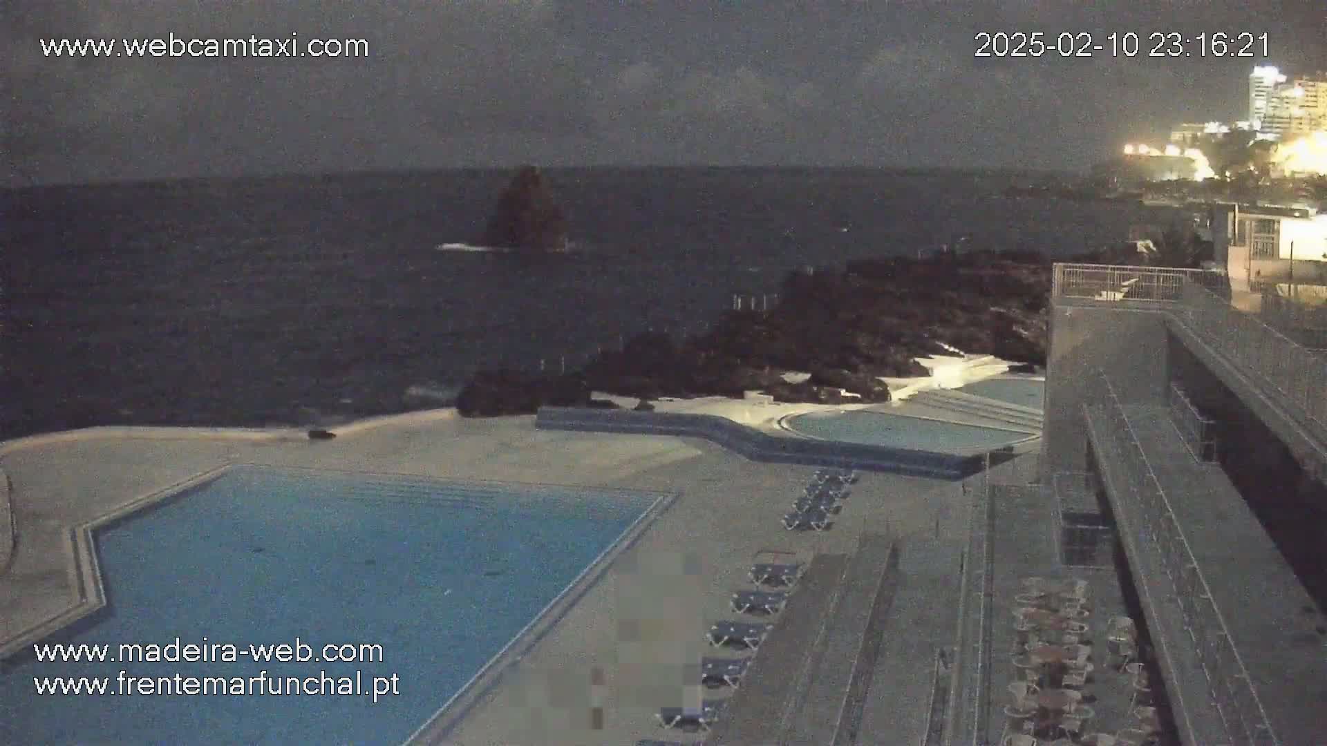 A nighttime view of an outdoor pool complex next to a rocky coastline with a dark, choppy ocean under a cloudy sky.