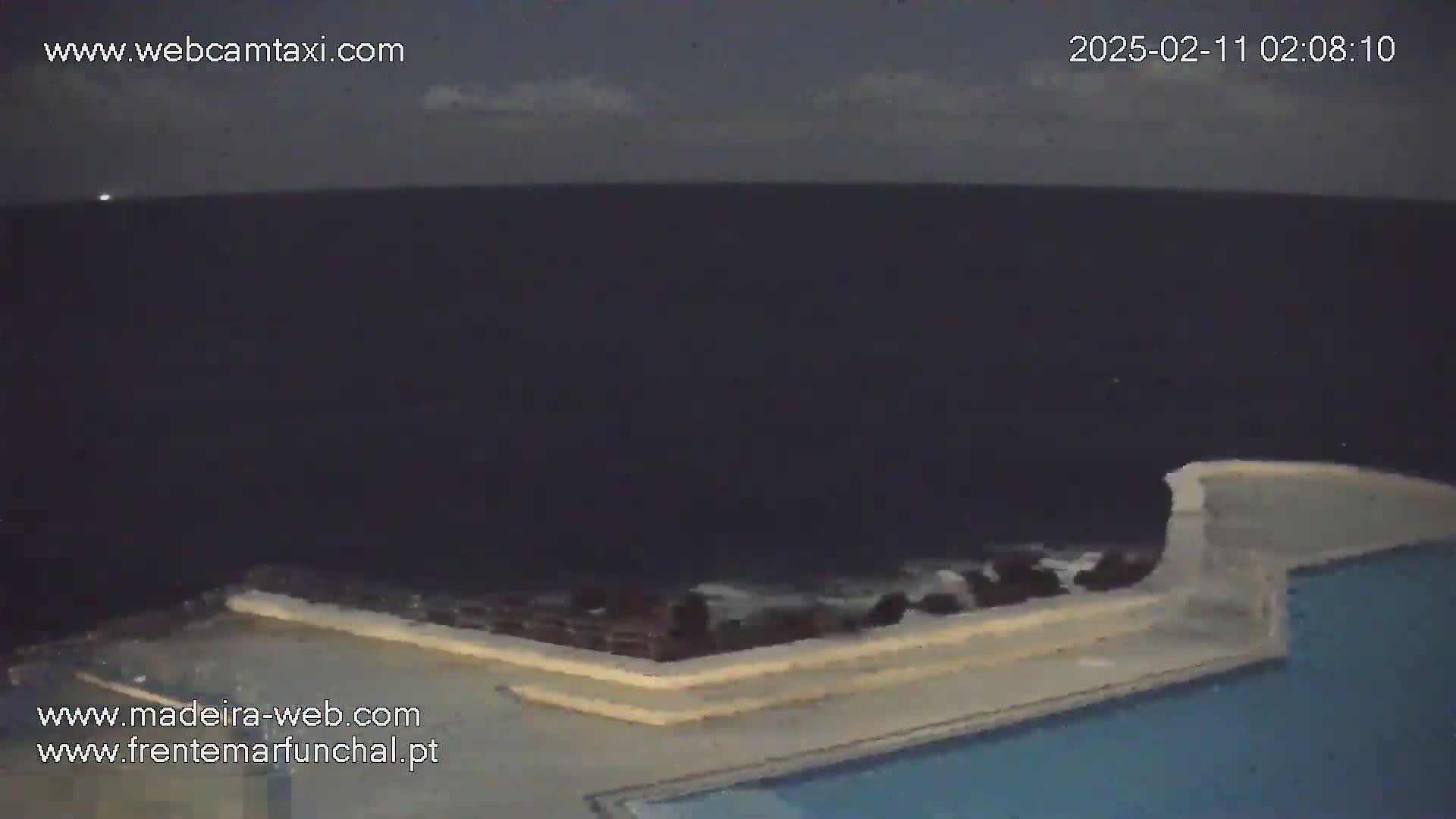 A nighttime view of a dark ocean under a mostly cloudy sky, partially visible above a low wall surrounding a dark blue pool.