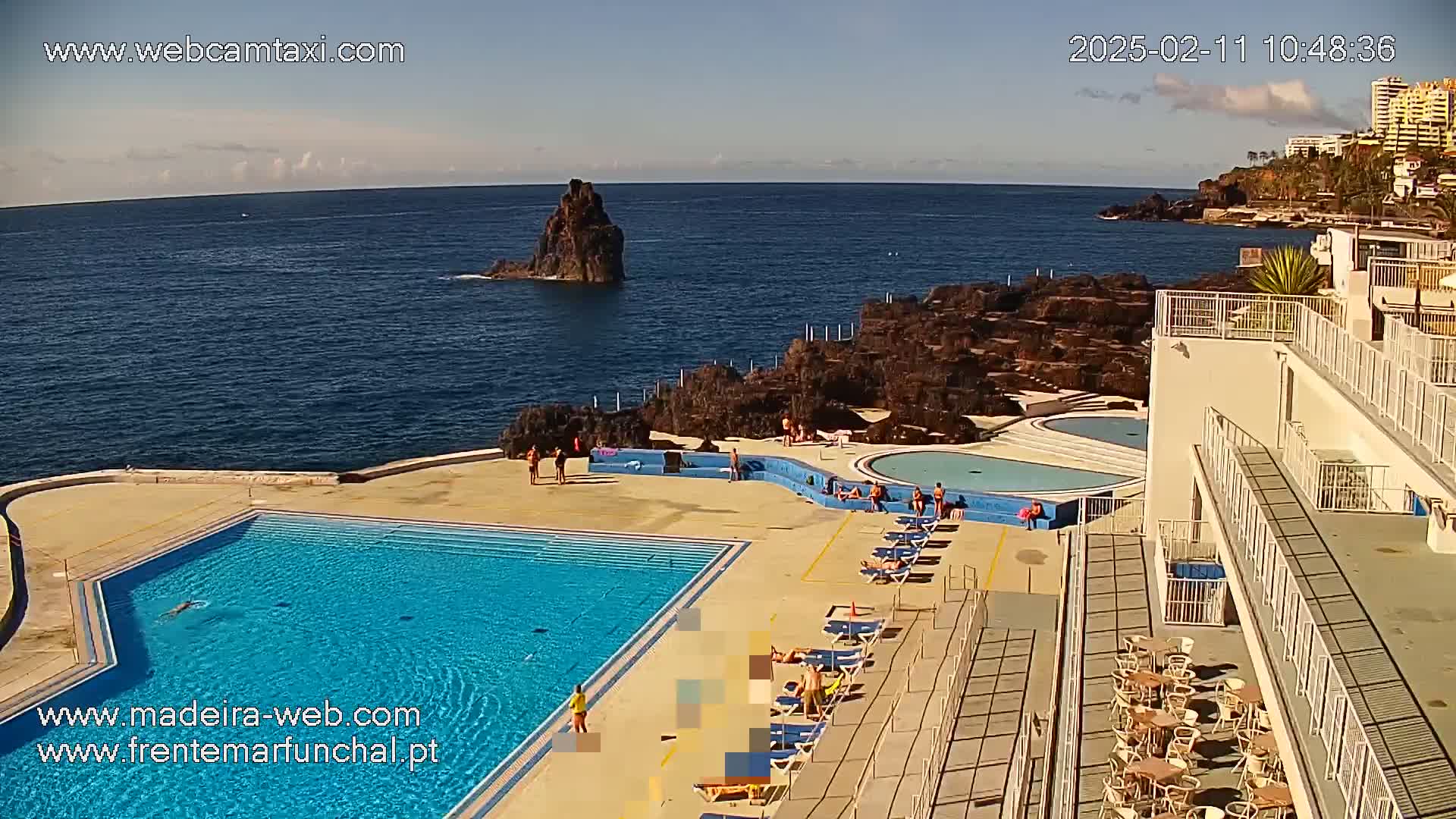 A swimming pool complex overlooks a rocky coastline and ocean under a mostly clear sky, with people relaxing in and around the pool area.