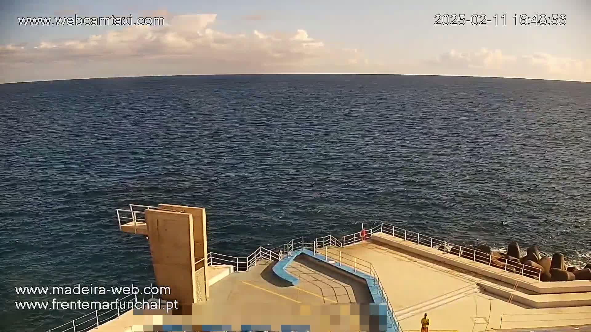 A mostly sunny day overlooking a dark blue ocean from a concrete platform with a diving board and breakwater.