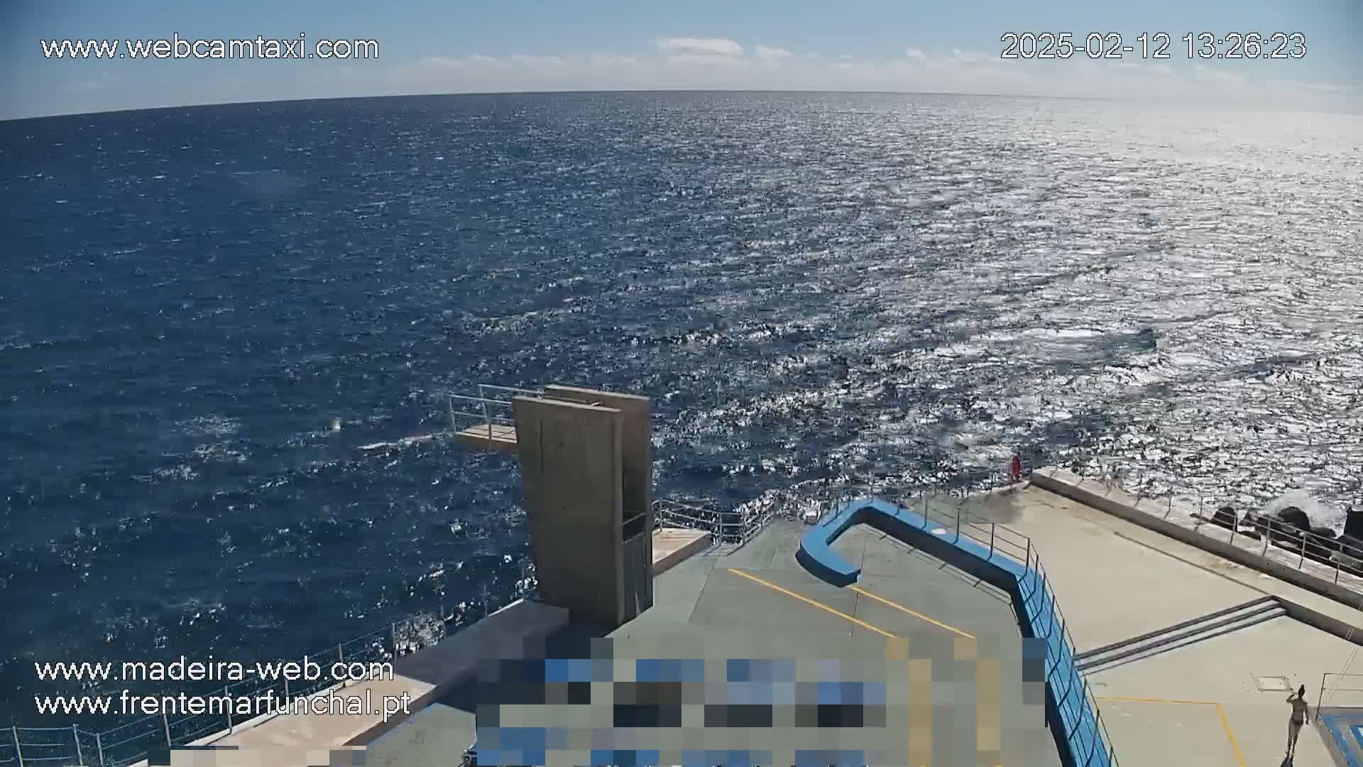 A diving platform overlooks a wavy, sunlit ocean on a partly cloudy day.