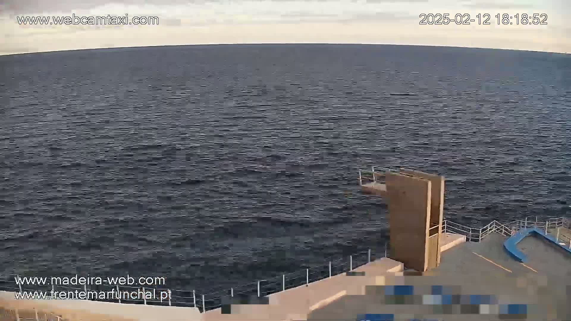 A mostly calm body of dark water is seen from an elevated vantage point, with a diving platform and surrounding concrete visible in the foreground under a slightly cloudy sky.