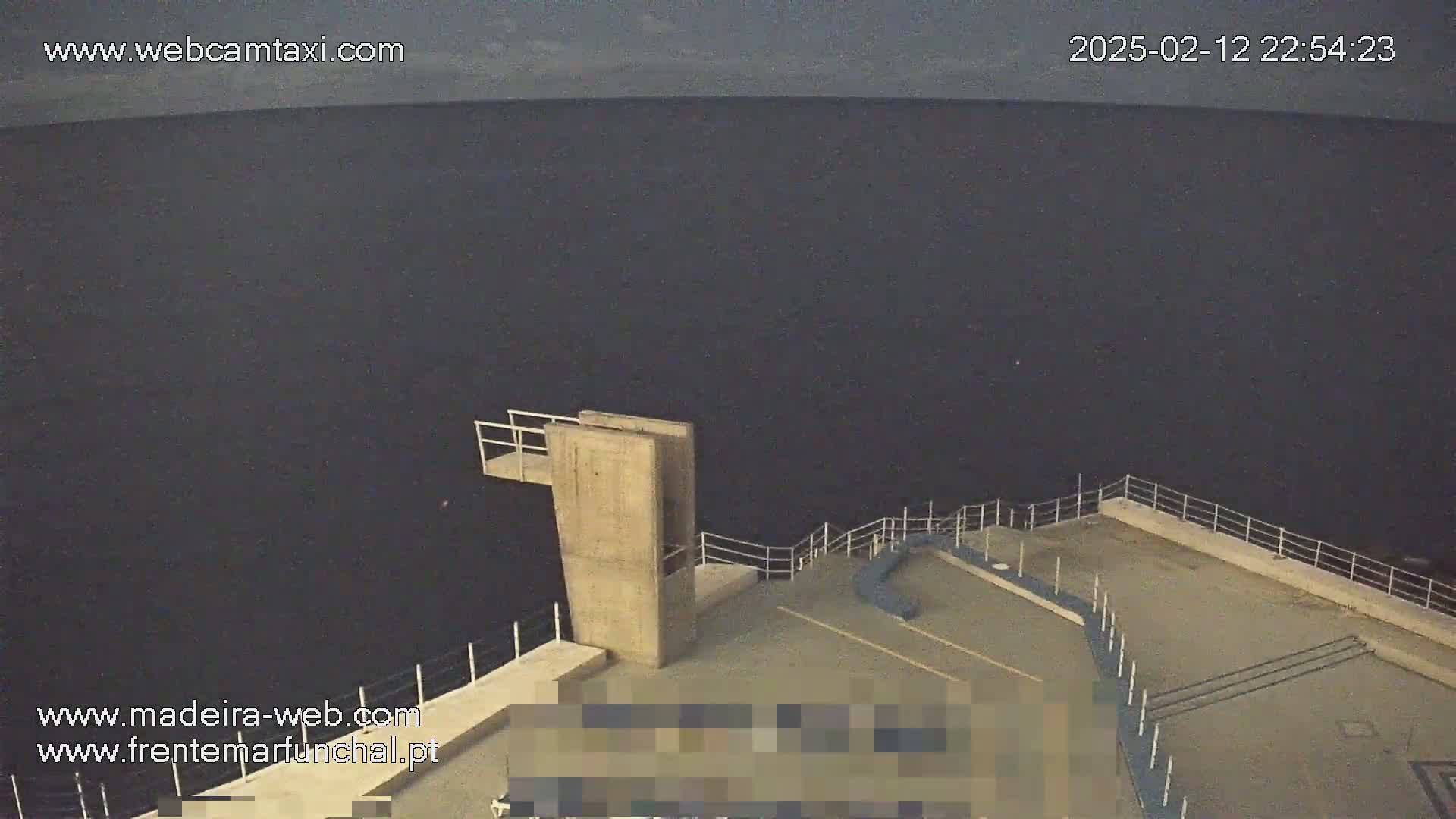 A high-angle, nighttime view shows a concrete diving platform overlooking a dark body of water under a cloudy sky.