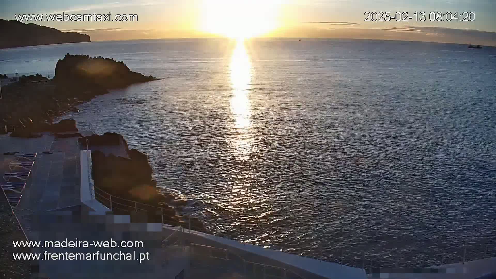 A sunrise over a calm ocean, illuminating the water with a bright path of light, next to a rocky shoreline with a concrete deck.