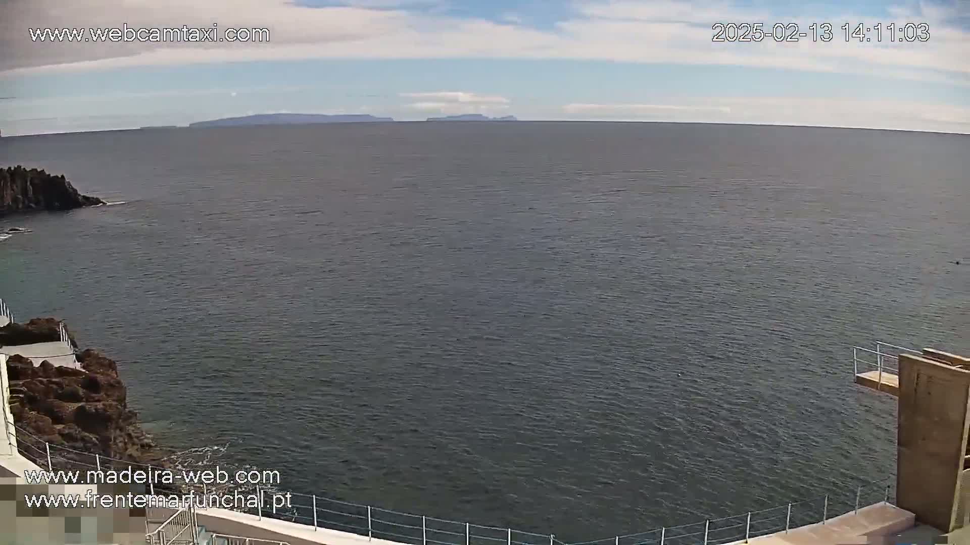 A mostly sunny day shows a wide expanse of dark-grey ocean meeting a rocky shoreline with a diving platform.