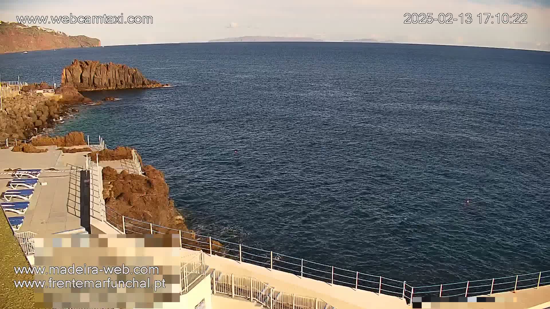 A rocky coastline with a paved area containing lounge chairs overlooks a calm, dark blue ocean under a mostly clear sky.