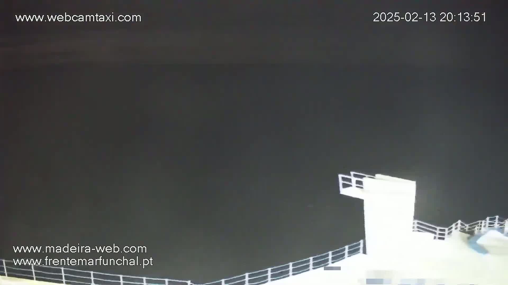 A nighttime view from a high vantage point shows a dark body of water and the upper portion of a white structure with railings, possibly a ship or dock.