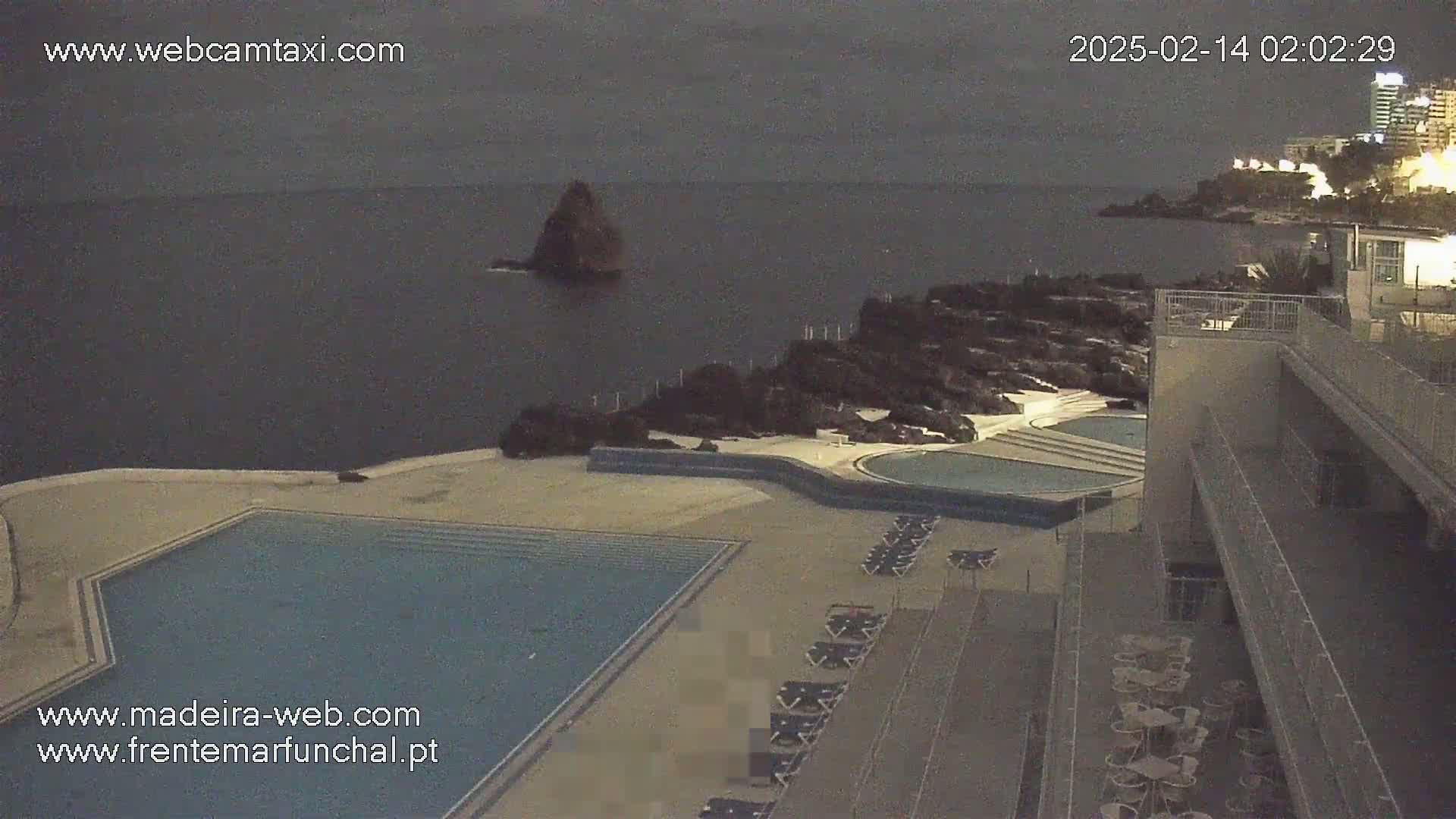 An outdoor nighttime scene shows a dark body of water, a rocky coastline, an empty swimming pool, and rows of empty lounge chairs next to a building.