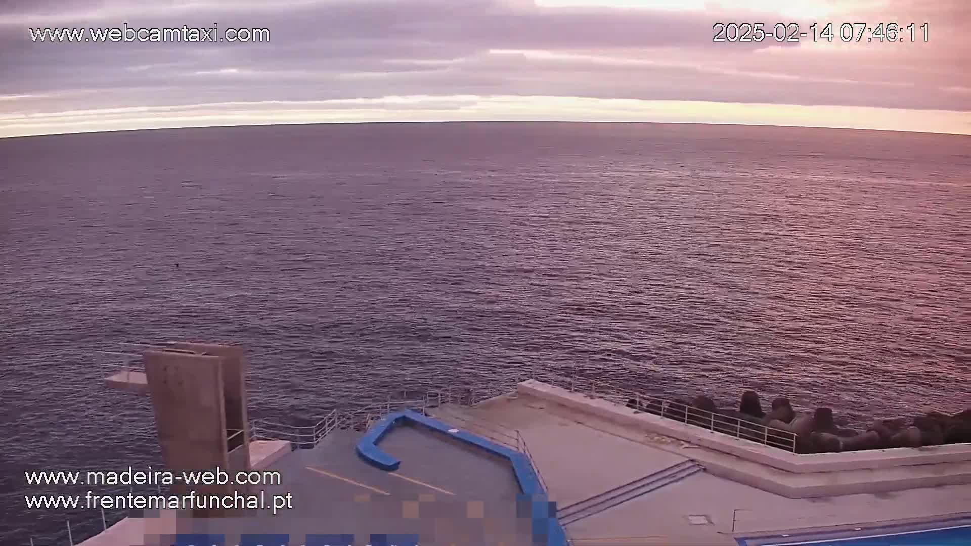 A calm, purplish-hued ocean stretches to the horizon under a cloudy, dusky sky, overlooking a concrete structure with a diving platform and a small pool.
