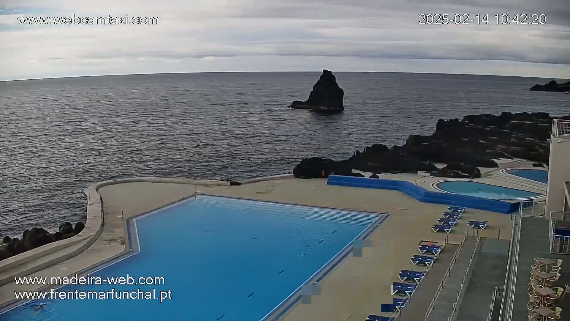 An outdoor swimming pool complex overlooks a dark-colored ocean under a cloudy sky, with a rocky outcrop visible in the distance.