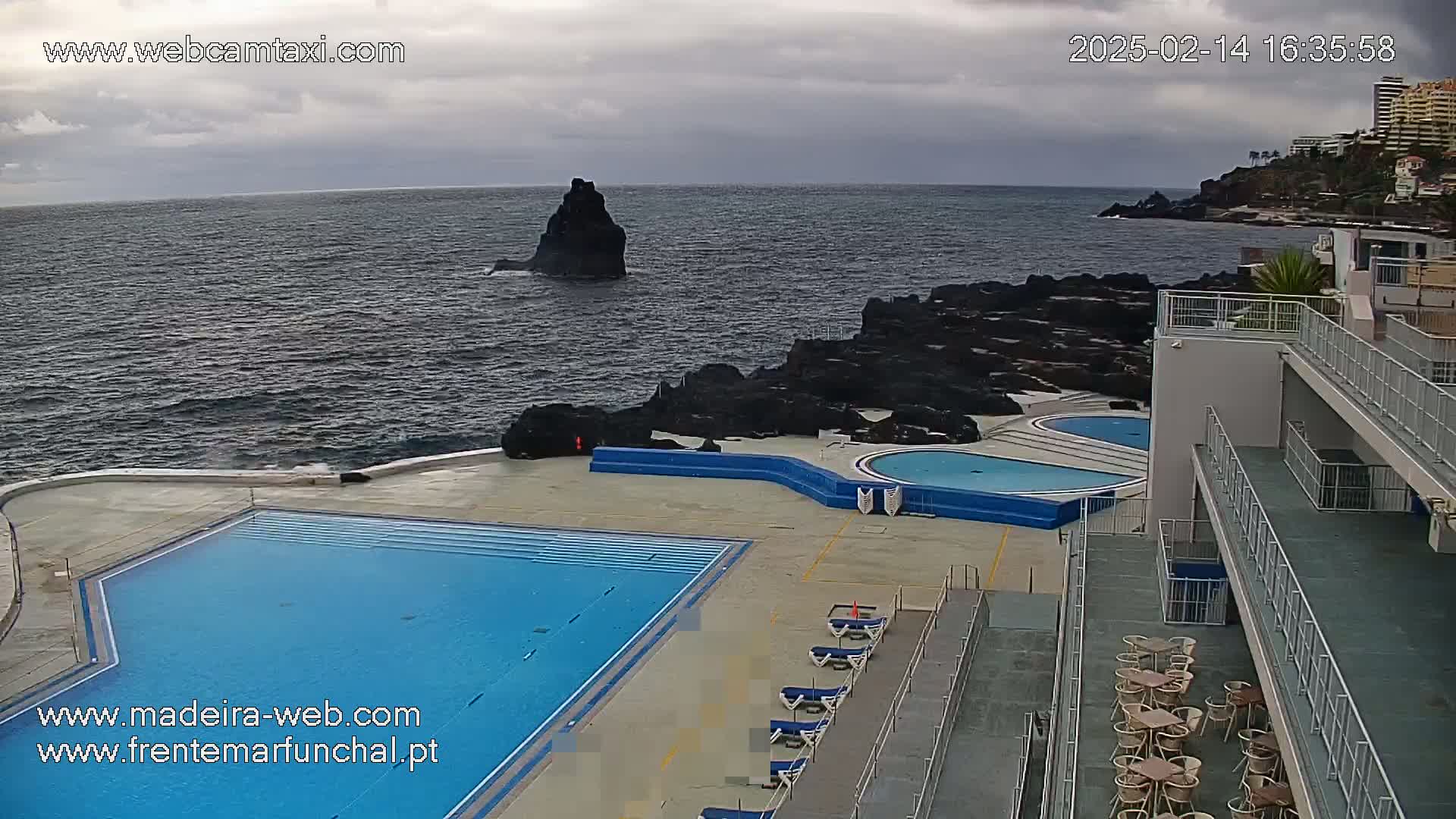 An outdoor pool complex overlooks a dark, wavy ocean under a cloudy sky, with a rocky shoreline and a distant rock formation visible.