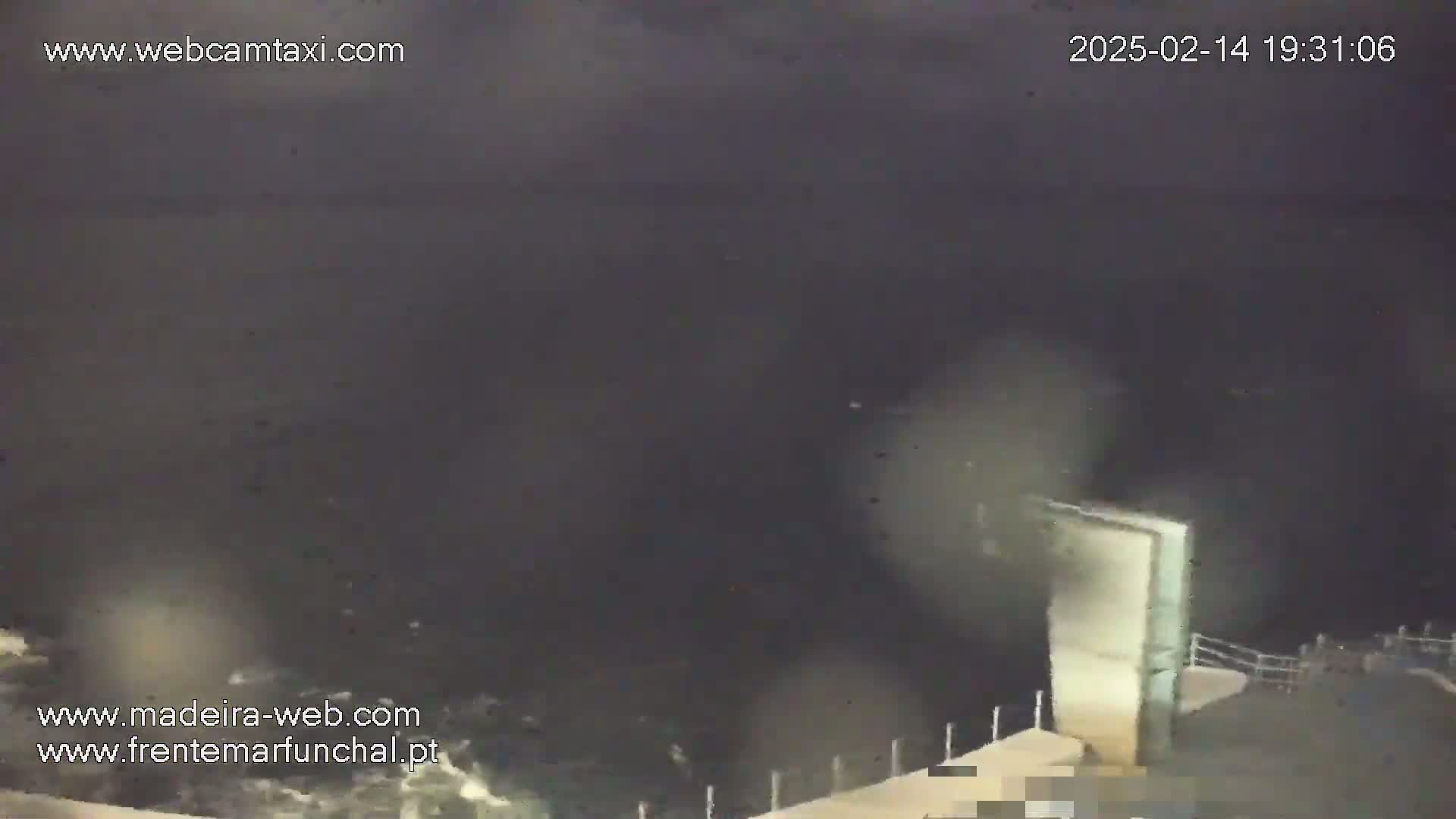 A nighttime, dark view of a rough sea crashing against a light-colored structure on a coast.