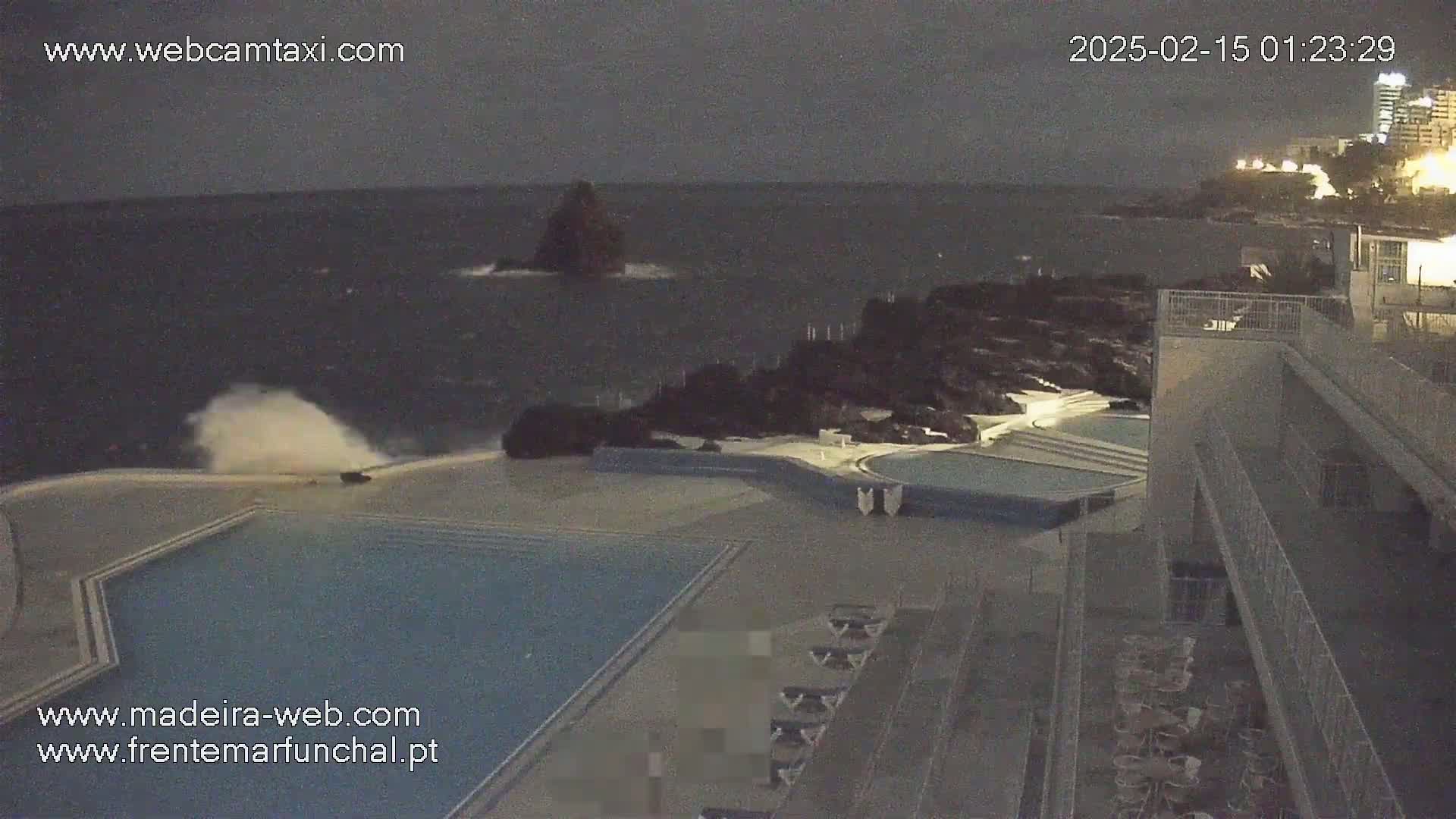 A nighttime view of a pool area next to a rocky coastline with a dark and turbulent ocean, under a cloudy sky.
