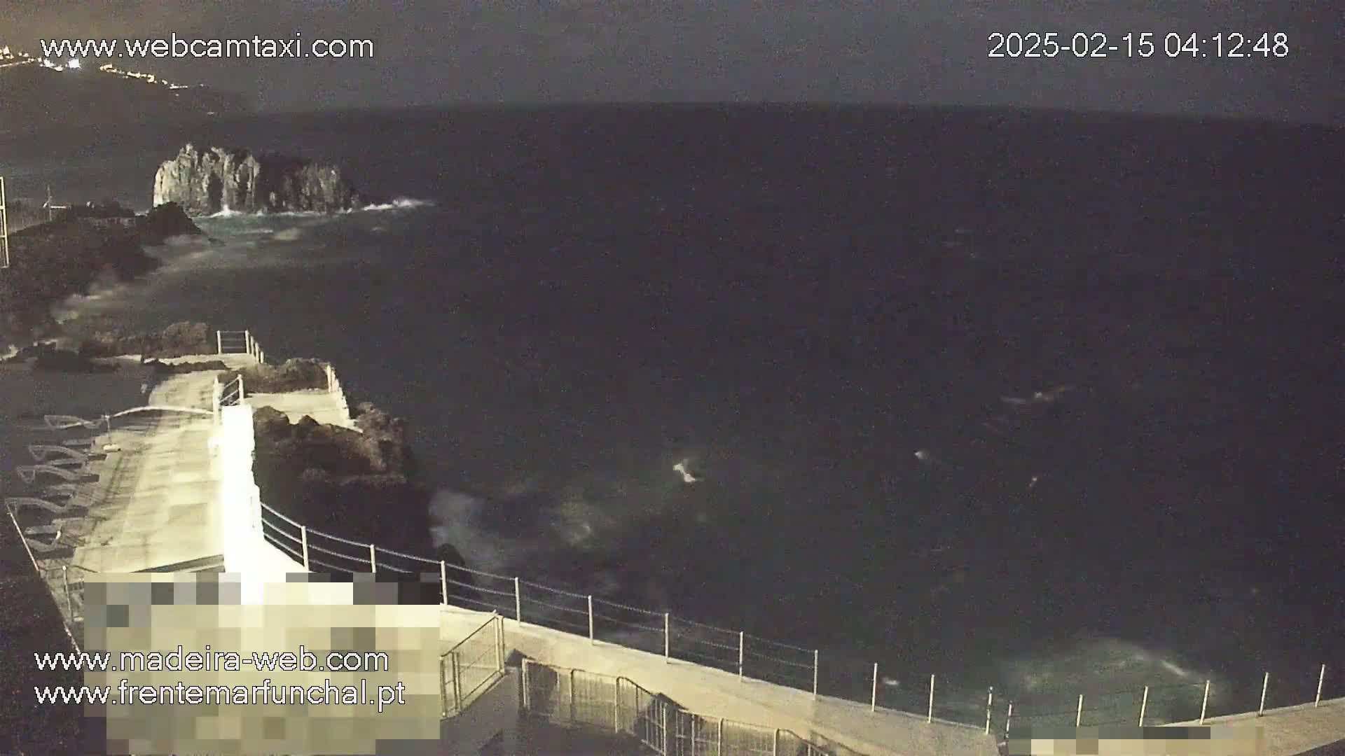 A nighttime coastal scene shows dark, choppy water crashing against a rocky shoreline with a concrete walkway and metal railing.